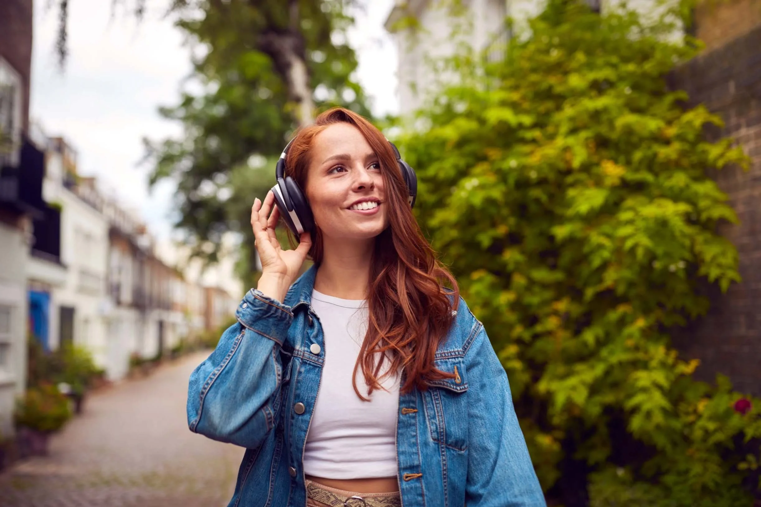 Woman wearing headphones and a denim jacket smiling as she walks down a street, product-in-use lifestyle photography in London by Alex Gell.