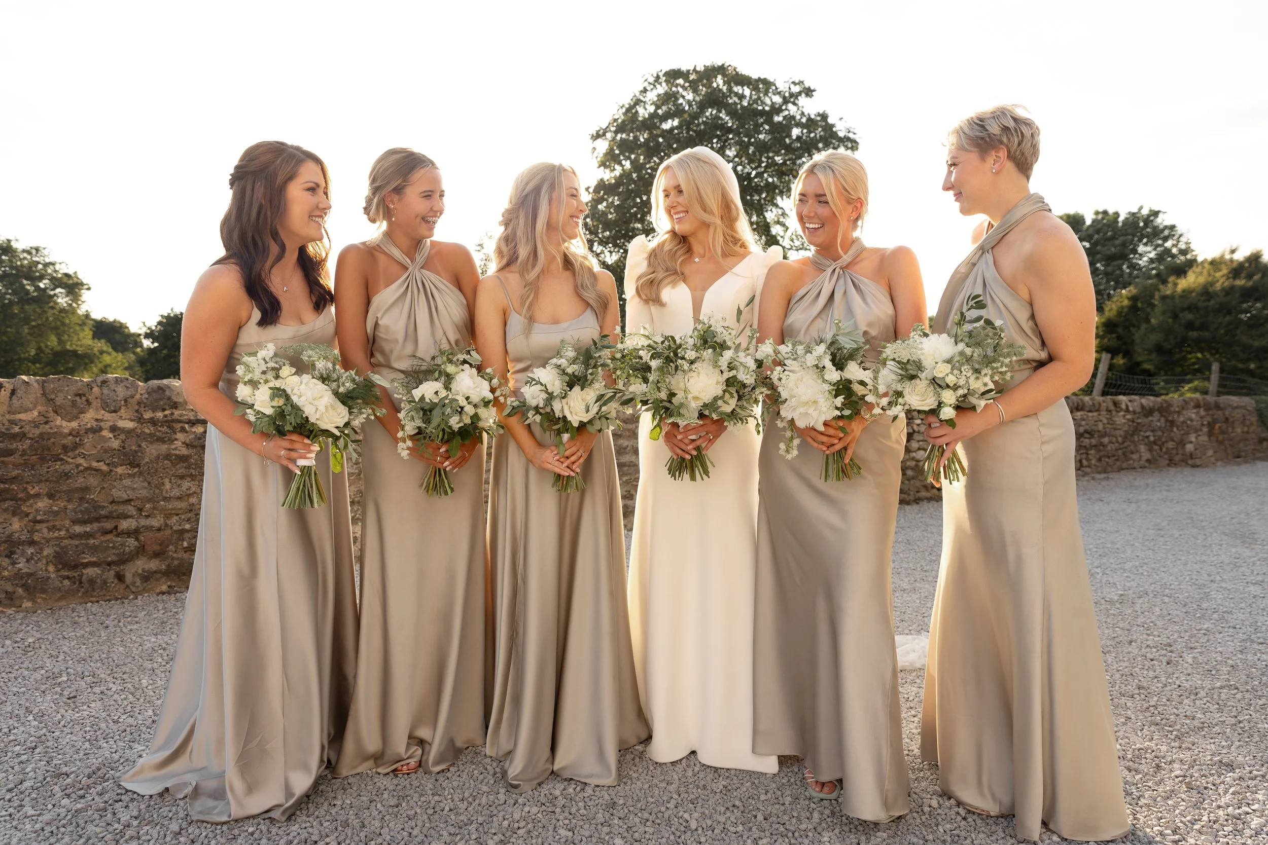 A group of six women dressed in matching beige dresses, holding bouquets of white flowers, standing outdoors during sunset.