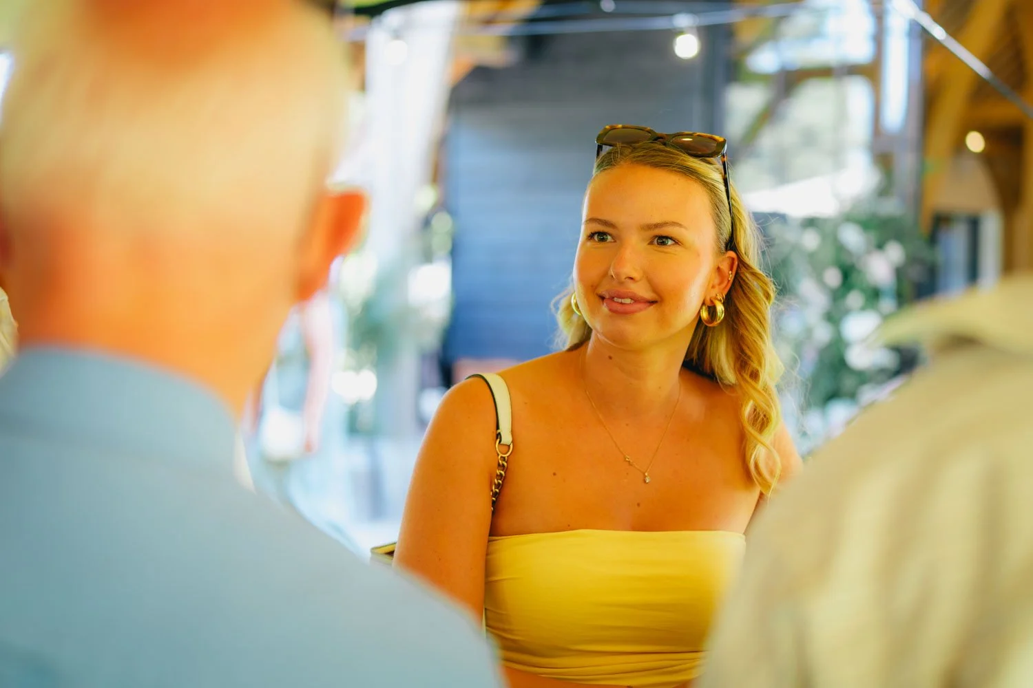 A woman with blonde hair wearing sunglasses on her head, gold hoop earrings, a yellow strapless top, and a gold necklace, smiling and engaging in conversation with people at an outdoor setting.