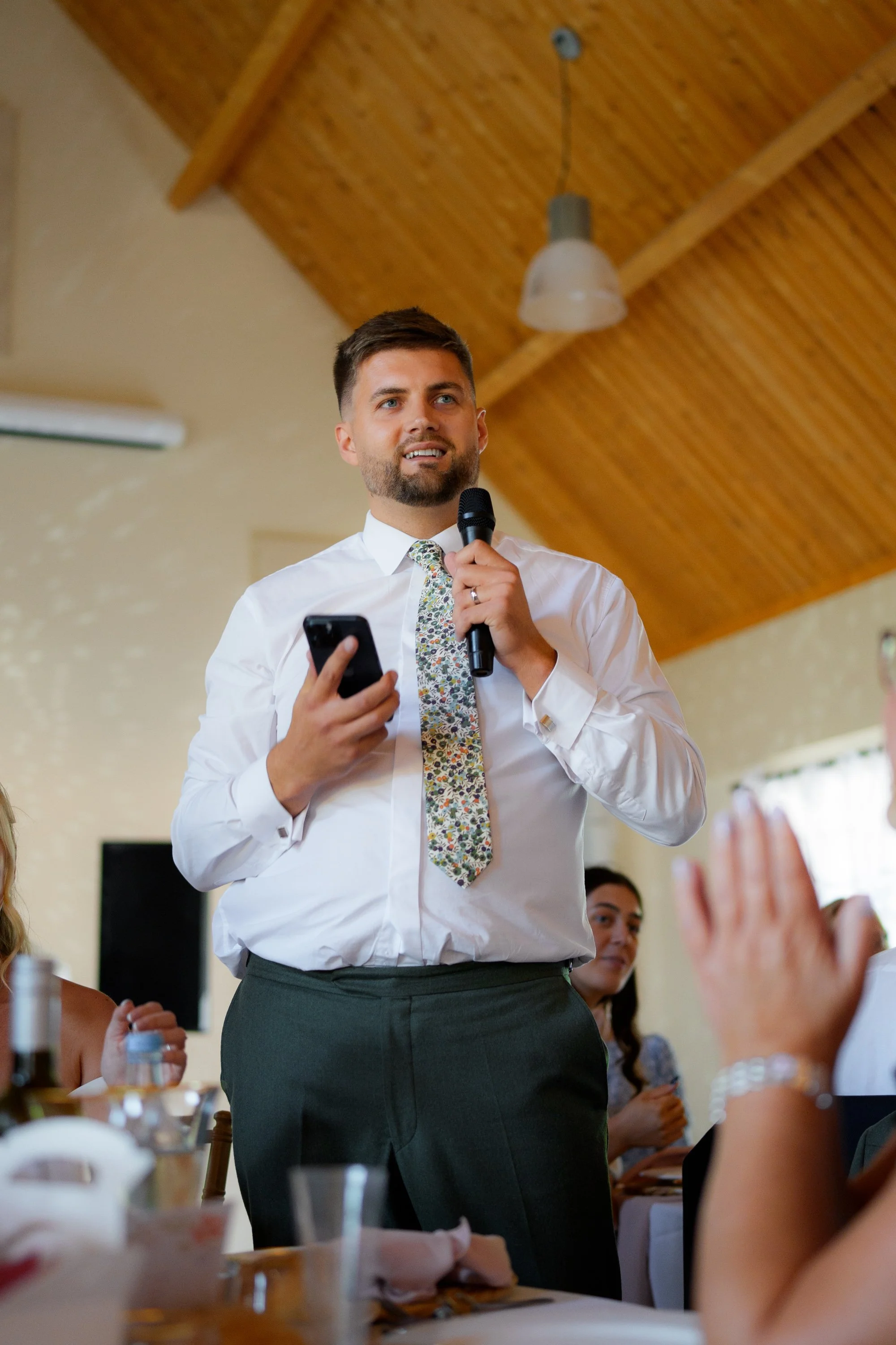 A man in a white shirt and colorful tie is speaking into a microphone during a gathering. Several people are seated around a table, raising their hands, and listening attentively.