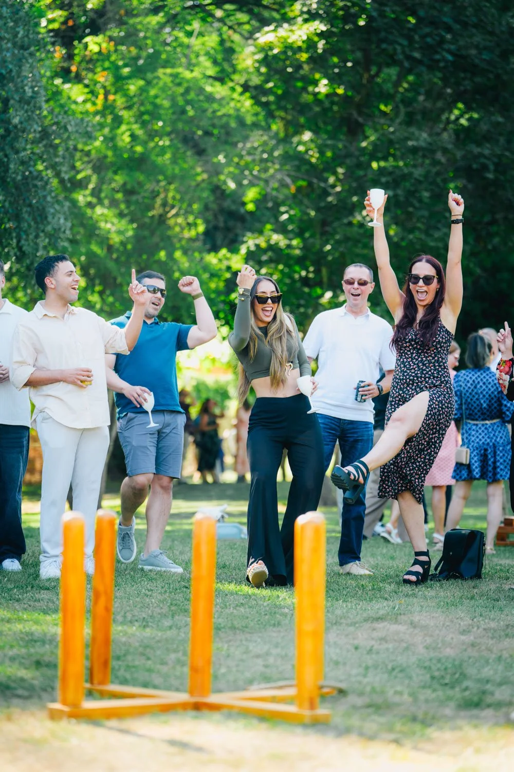 Group of friends celebrating outdoors, playing a game with small orange stakes, some holding drinks, smiling, and raising their hands.