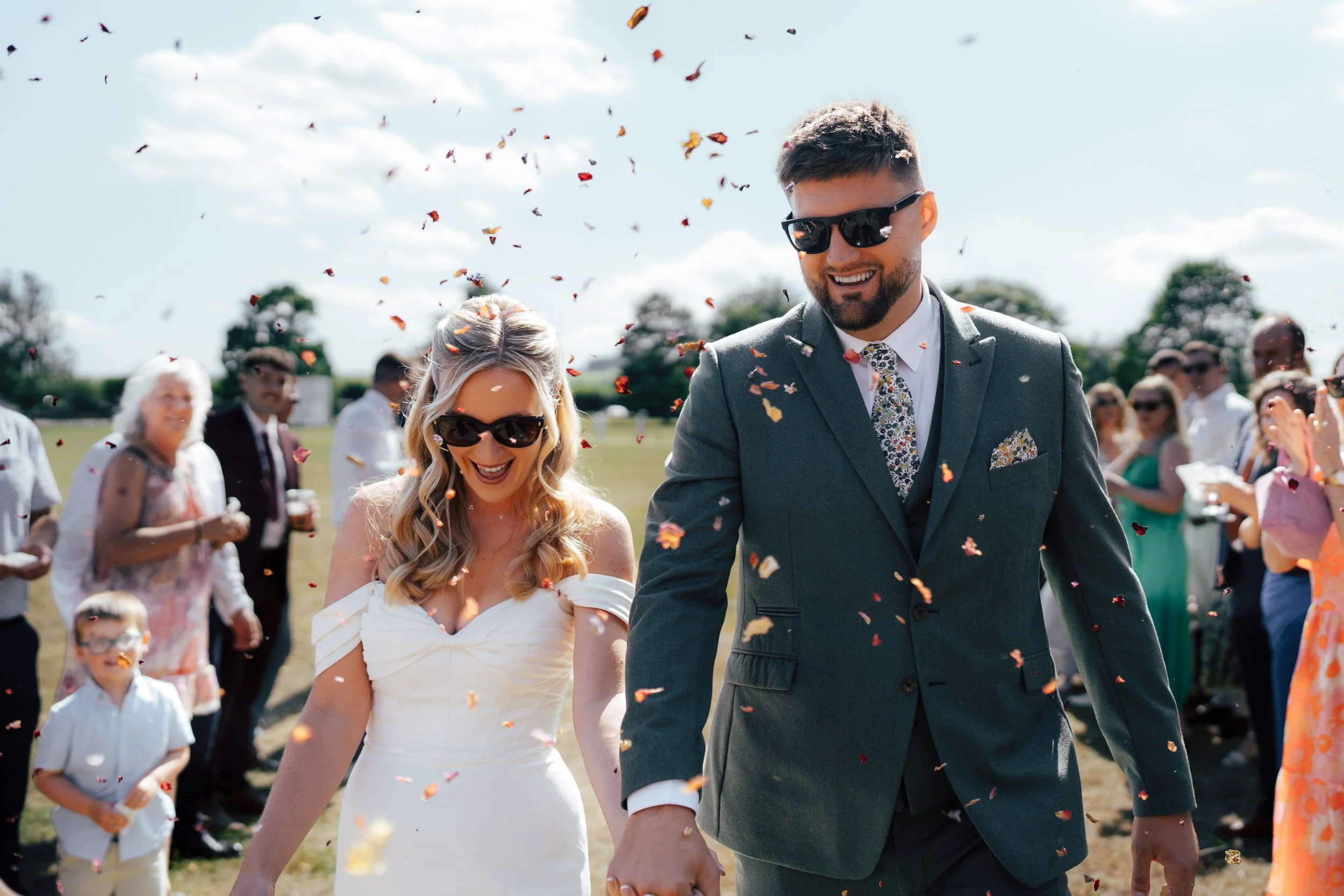 A newlywed couple, a woman in a white wedding dress with blonde hair and sunglasses, and a man in a dark suit with a floral tie and sunglasses, are smiling and holding hands as they walk through falling flower petals at an outdoor wedding celebration