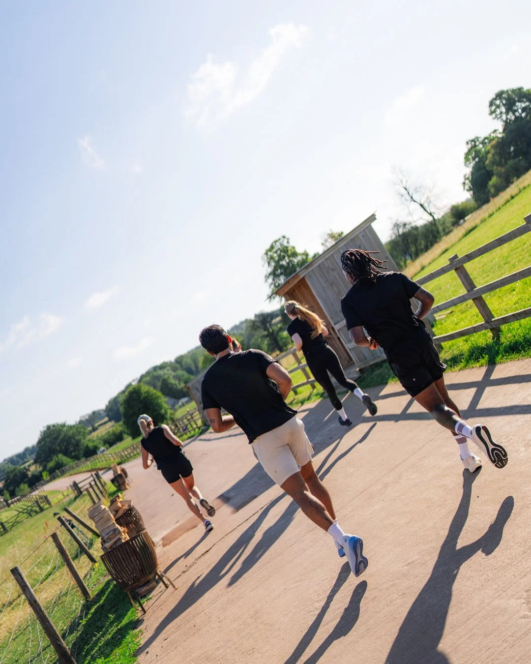 A group of five people running outdoors on a sunny day along a paved path with green fields and trees in the background.