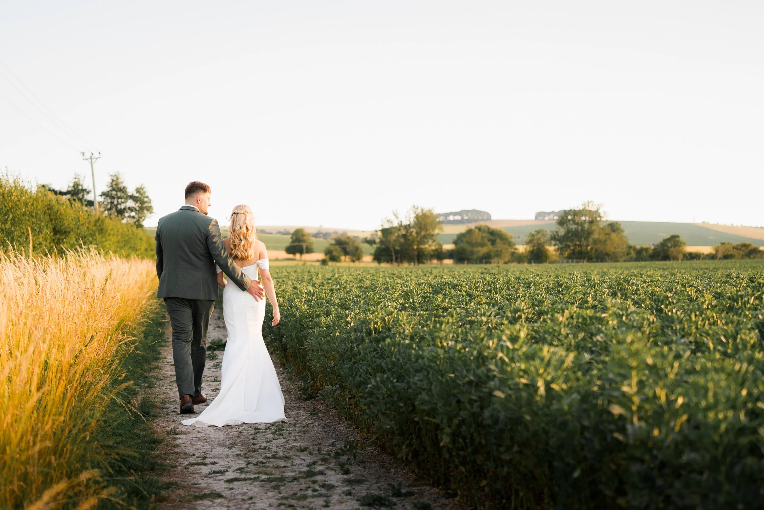 A bride and groom walking together through a farm field at sunset, holding hands. The groom is wearing a gray suit, and the bride is in a white wedding dress. The landscape features green fields, trees, and distant hills under a clear sky.