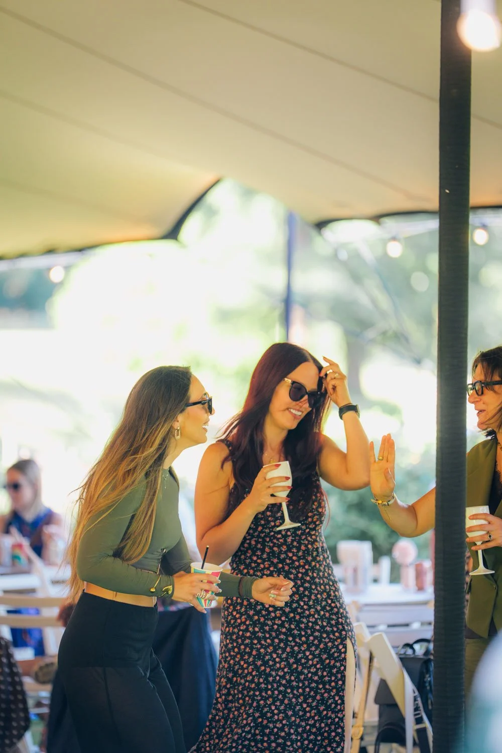 Three women wearing sunglasses and holding drinks are dancing and socializing under a tent with string lights at an outdoor gathering or party.