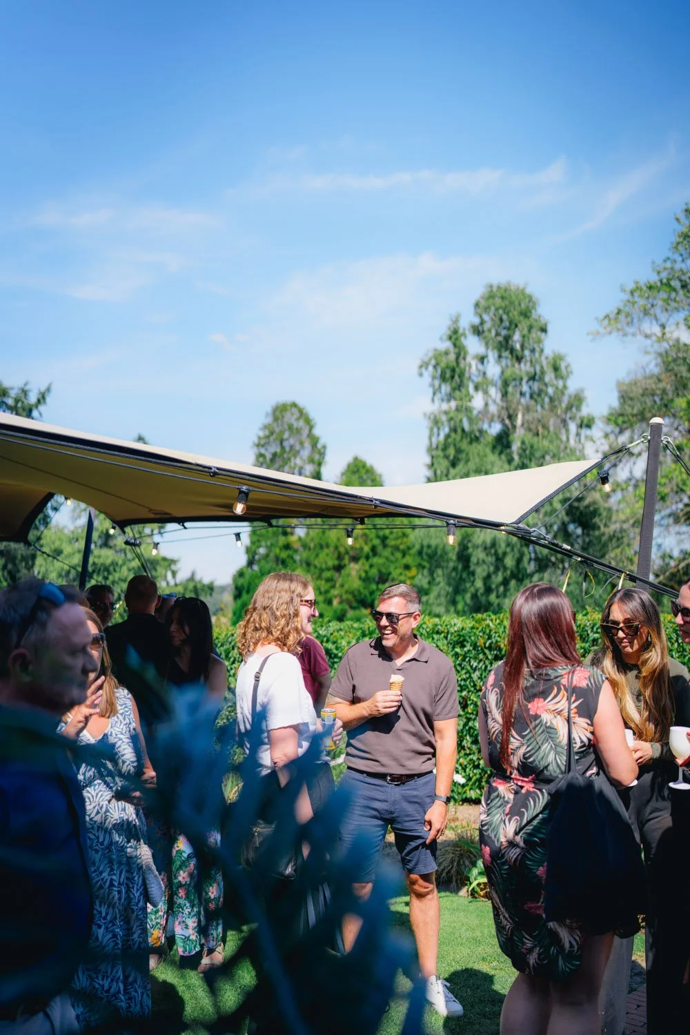People gathered outdoors on a sunny day, socializing under a canopy with string lights, with trees and greenery in the background.