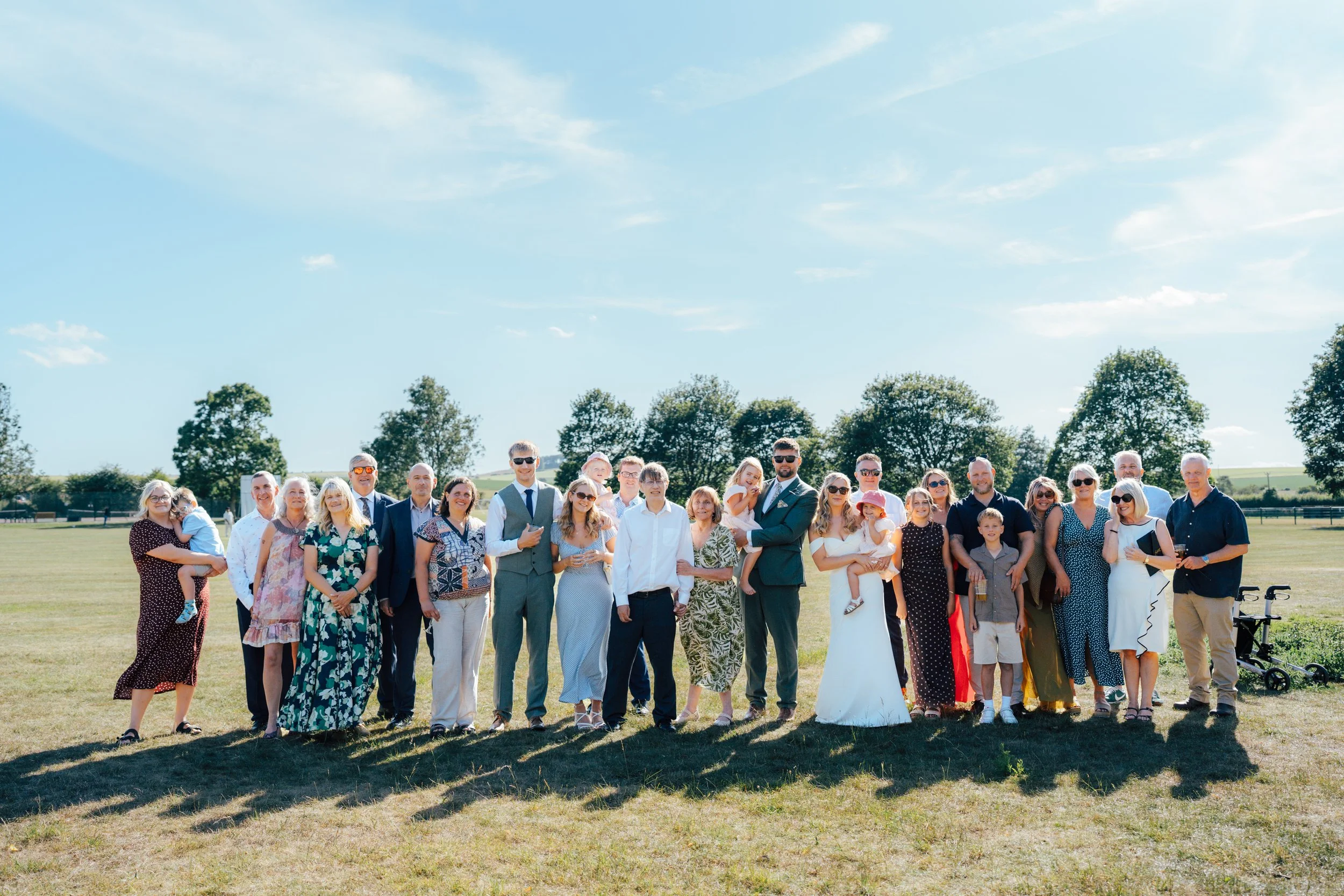 A large group of people gathered outdoors on a sunny day, standing on grass with trees in the background, posing for a photo.