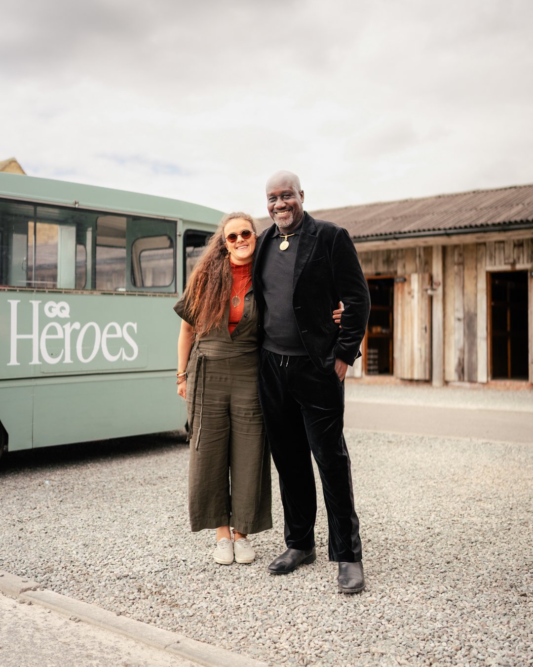 A smiling woman with long curly hair and sunglasses standing next to a tall man with a shaved head and beard, both dressed in casual stylish clothing, embracing outdoors in front of a vintage bus with the words 'GG Heroes' on it and a rustic wooden b