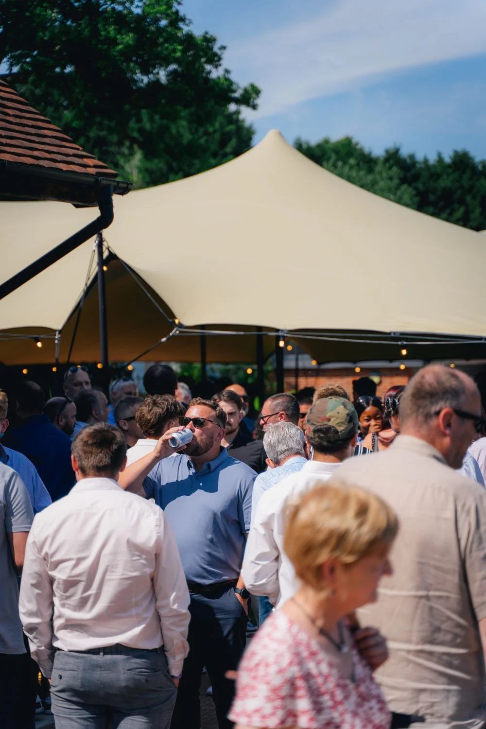A crowd of people gathered under a large beige patio umbrella outdoors during daytime, some are talking, some are drinking, and some are standing, with trees and a blue sky in the background.