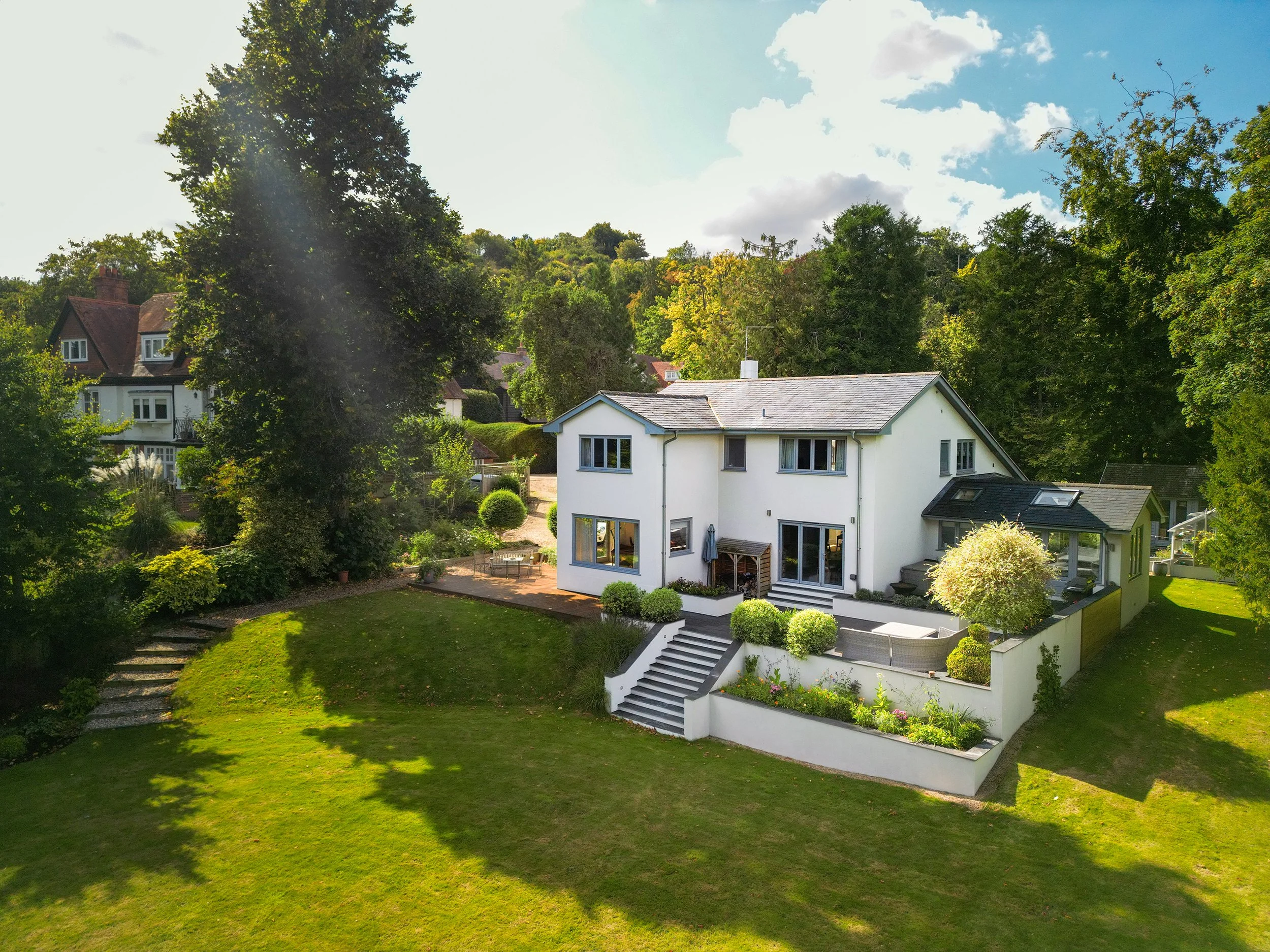 A white two-story house with a gray tiled roof, surrounded by green trees and a well-maintained lawn, with a landscaped garden and patio area.