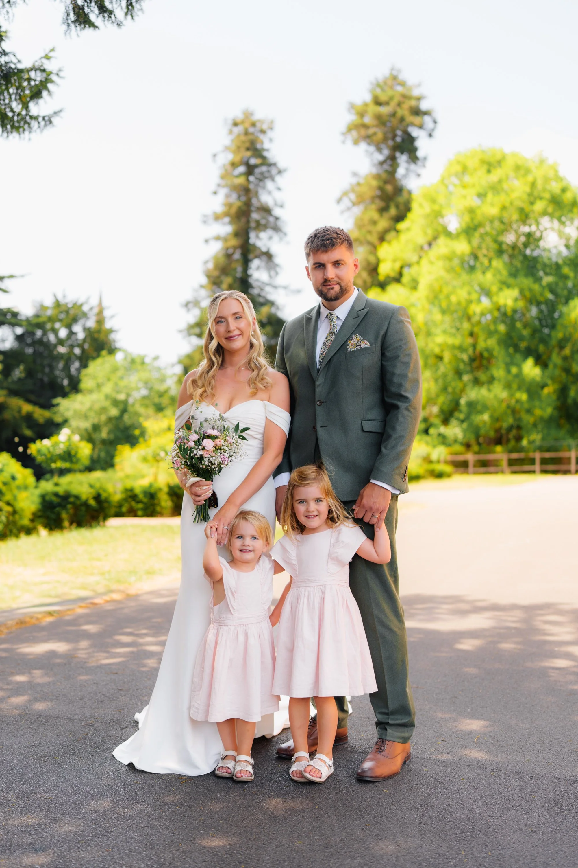 A wedding party in a park with a bride, groom, and three young girls, all smiling and dressed in formal attire, with lush green trees in the background.