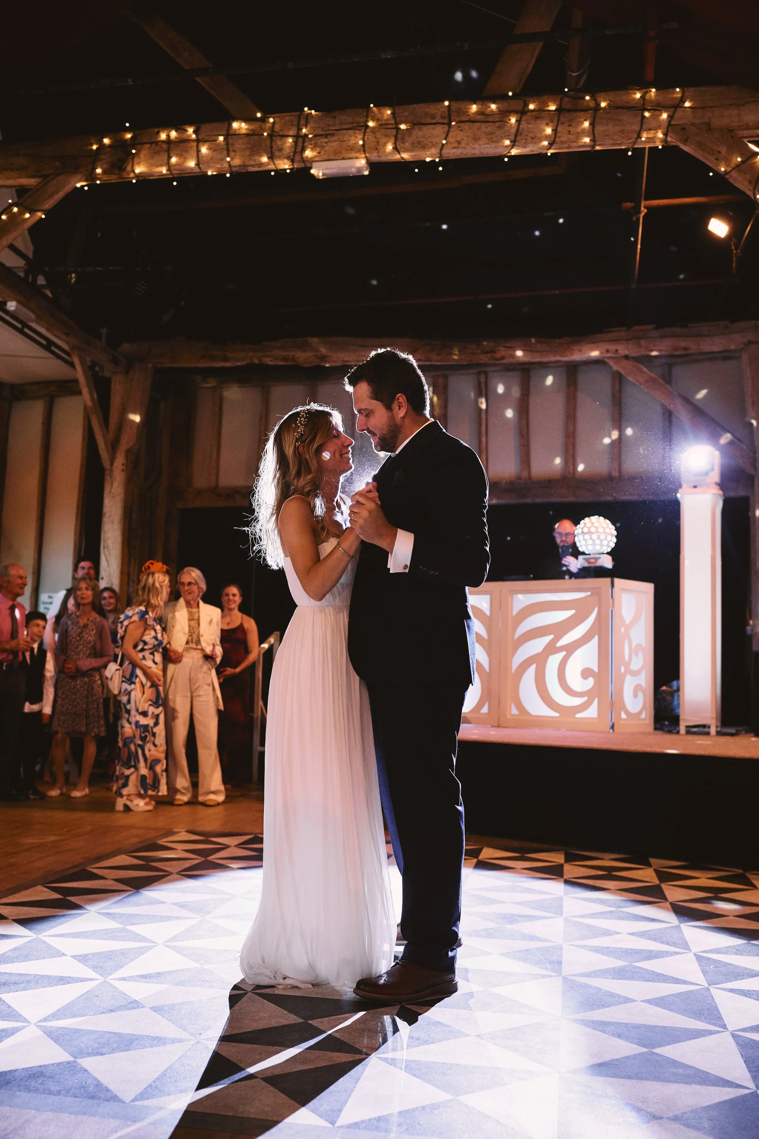 Couple dancing at their wedding reception in a rustic barn with string lights, surrounded by friends and family, with a DJ booth in the background.