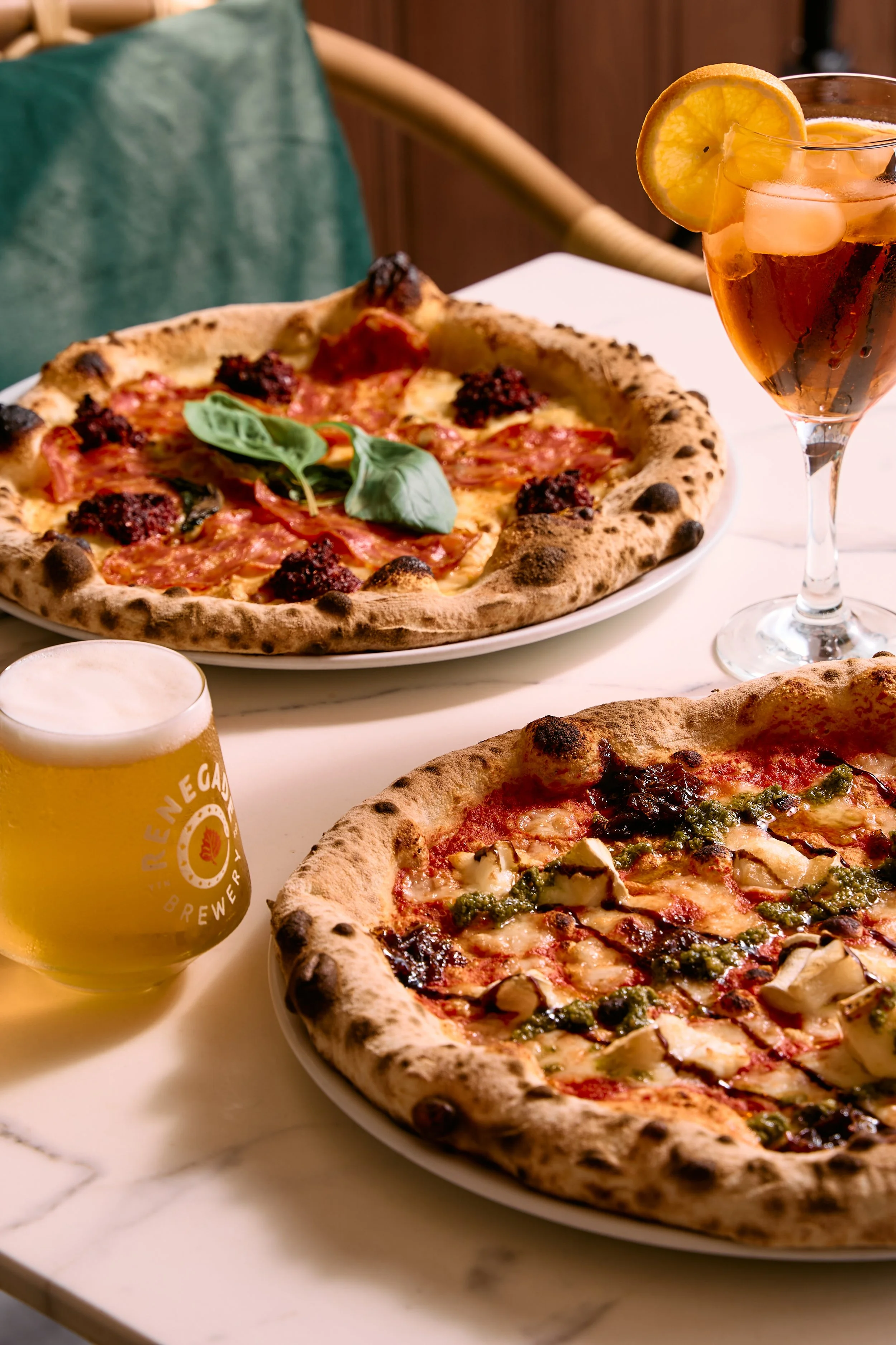 Two pizzas on white plates, one with pepperoni, basil, and dark toppings, the other with cheese, chicken, and green herbs, along with a glass of orange-colored cocktail with lemon slices and ice, and a glass of beer on a white table.