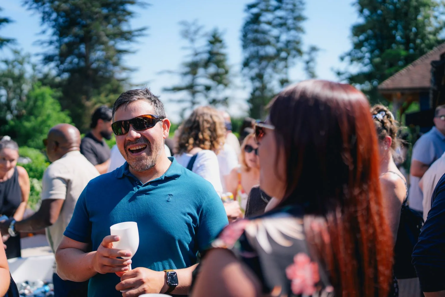 People gathered outdoors at a social event, smiling and chatting, with trees and a building in the background.