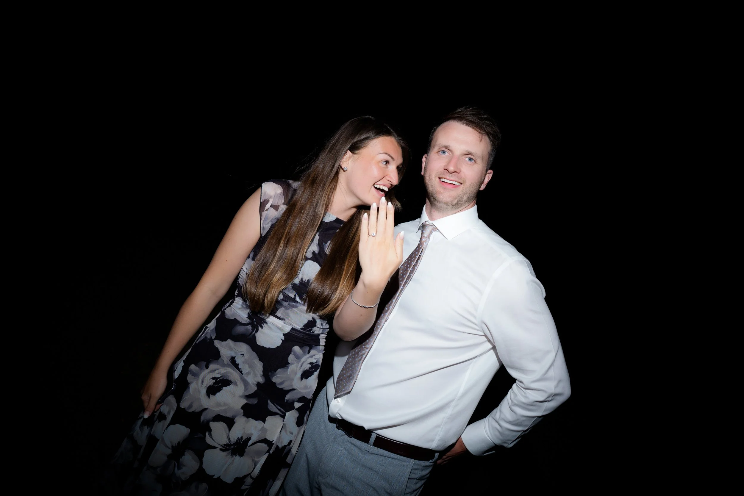 A woman and a man pose together against a black background, smiling with the woman whispering into the man's ear and displaying an engagement ring.