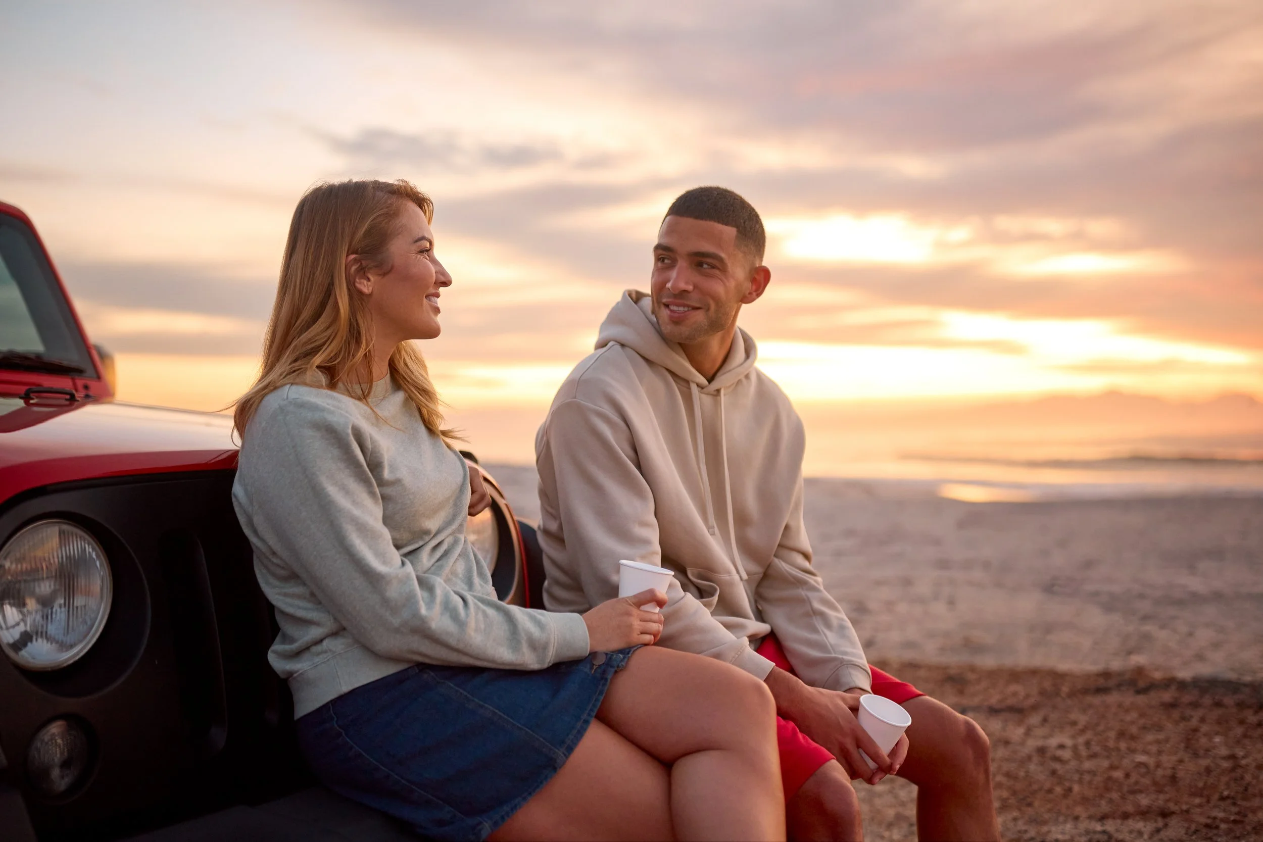 A man and a woman sitting on the hood of a red car at the beach during sunset, holding cups and chatting.