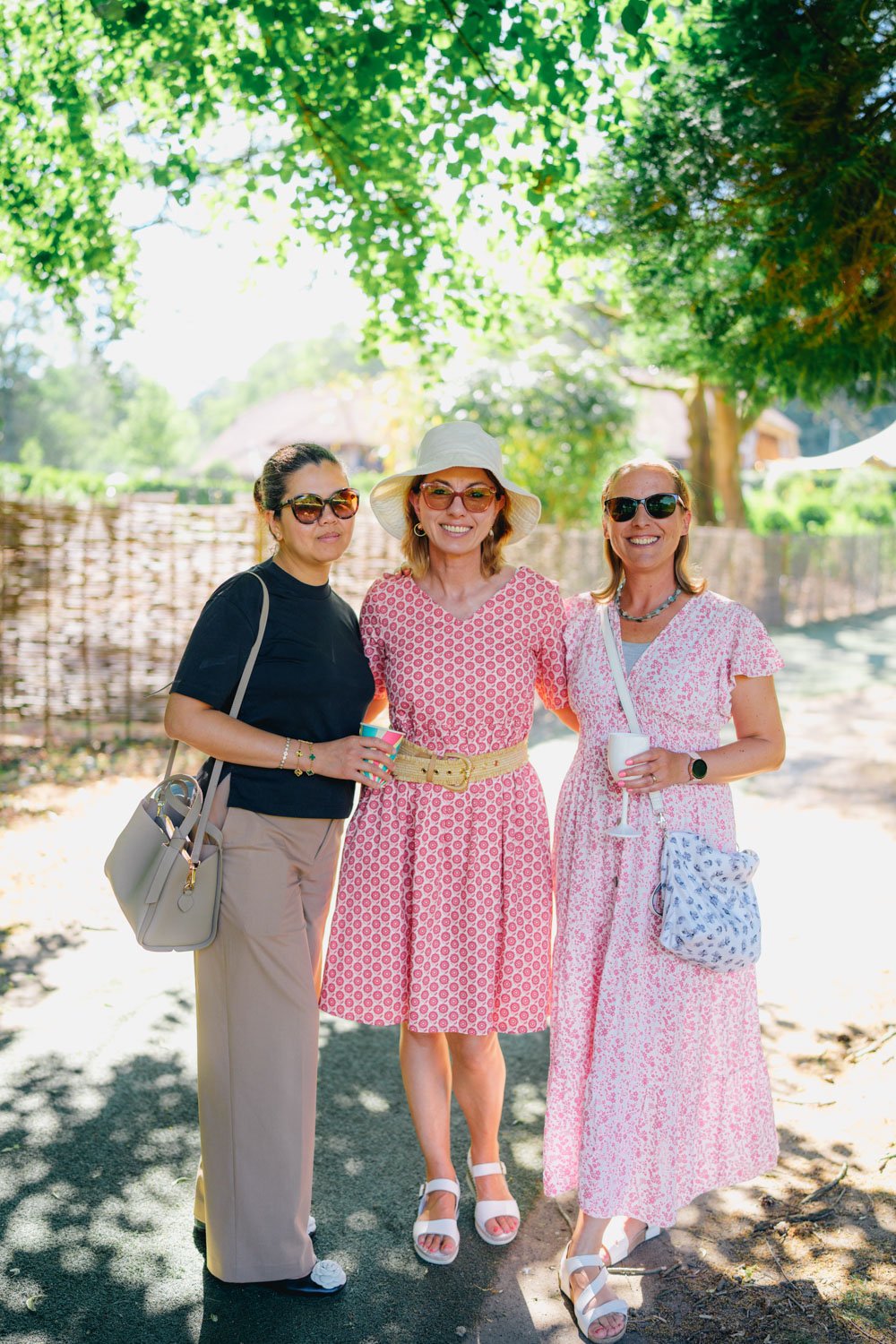 Three women standing outdoors in bright sunlight, smiling, wearing sunglasses, and dressed in summer outfits, with green trees and a fence in the background.
