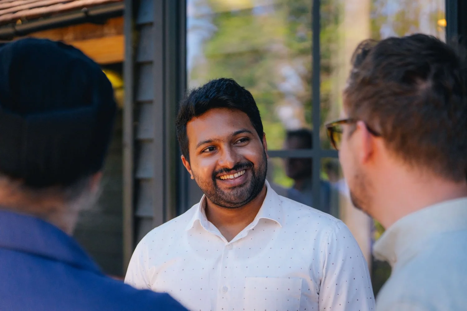 Three men having a conversation outdoors, smiling, with a background of trees and windows.