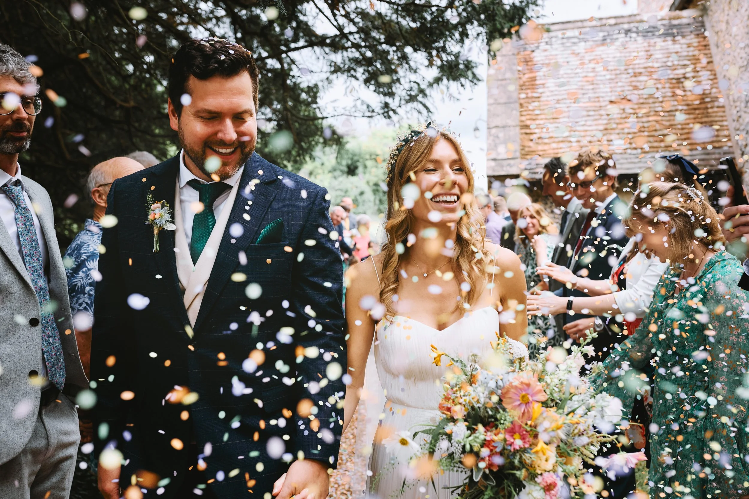 A smiling bride and groom holding hands surrounded by guests. Confetti falls around them at an outdoor wedding under trees, with a rustic building in the background.