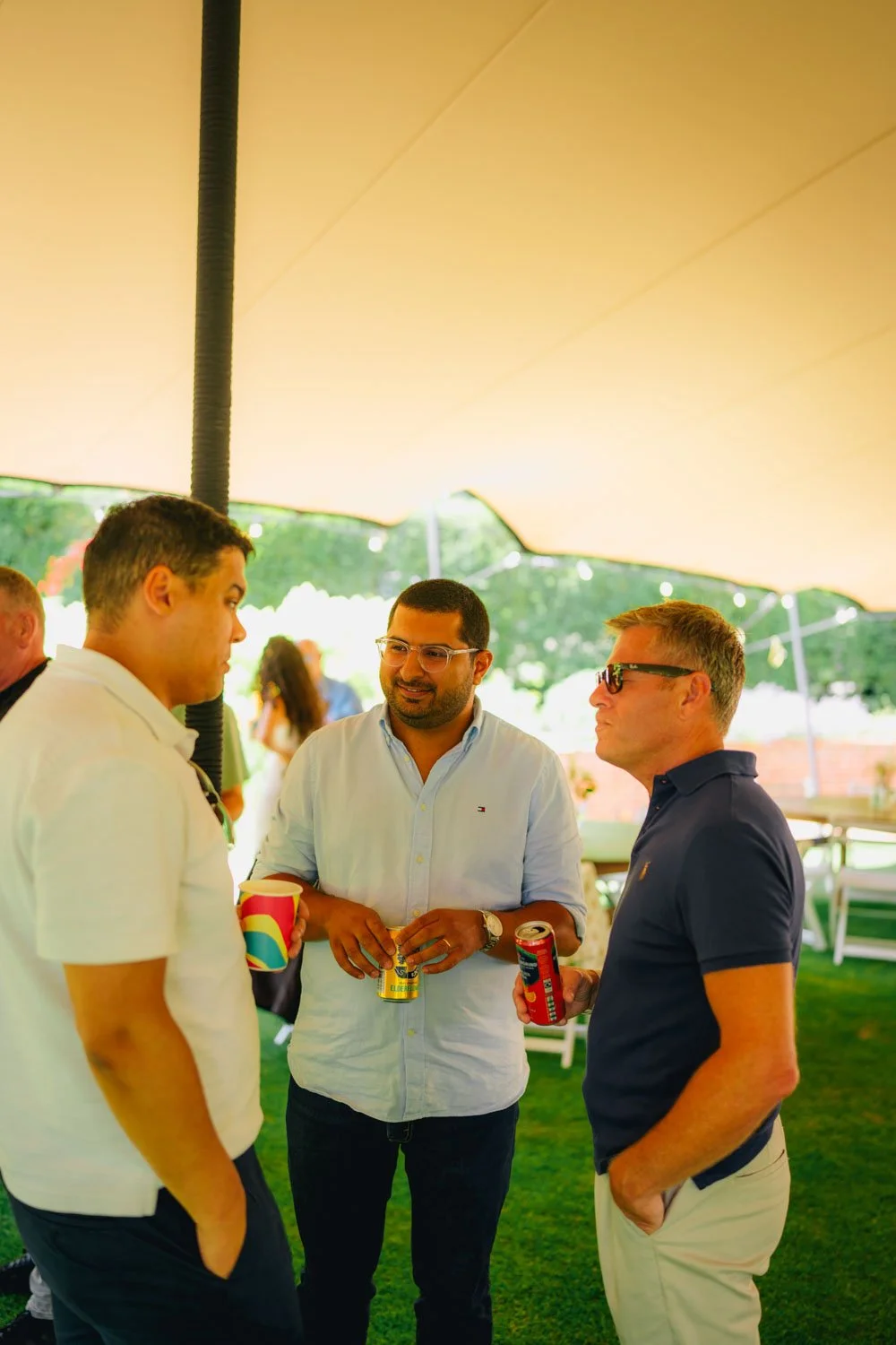 Three men talking at an outdoor event under a large beige canopy. They are holding canned drinks and are casually dressed, with two wearing sunglasses and one with clear glasses. There are tables and chairs in the background, with greenery visible be
