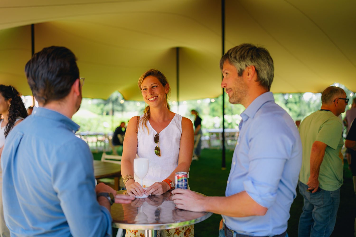 Three people are standing around a high-top table outdoors at a social event, smiling and drinking. Two men wear light blue shirts, and a woman wears a sleeveless white top with sunglasses hanging from her shirt. There are drinks on the table, and ot