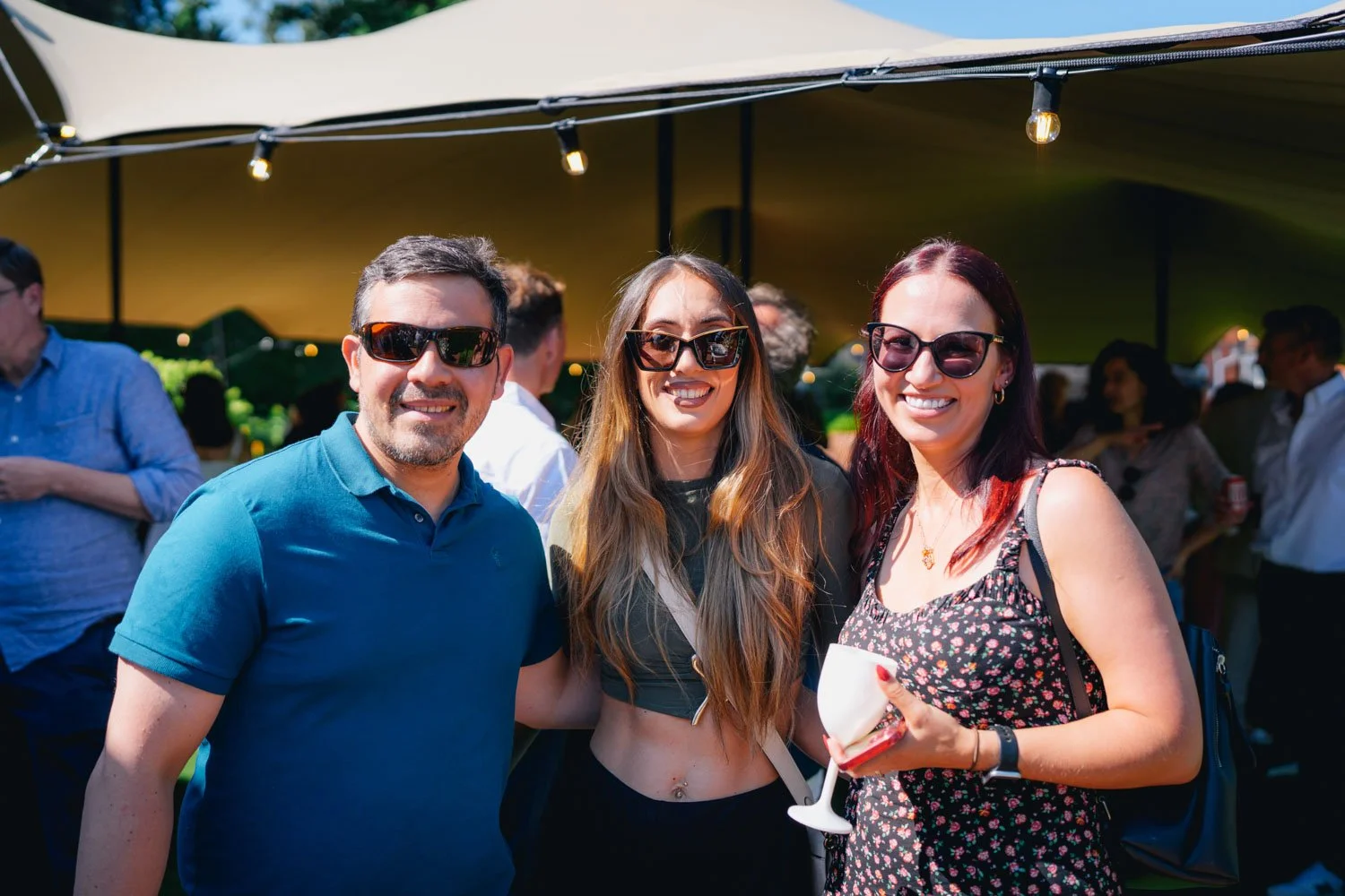 Three friends wearing sunglasses smiling at a social outdoor gathering under a large canopy, with other people in the background.