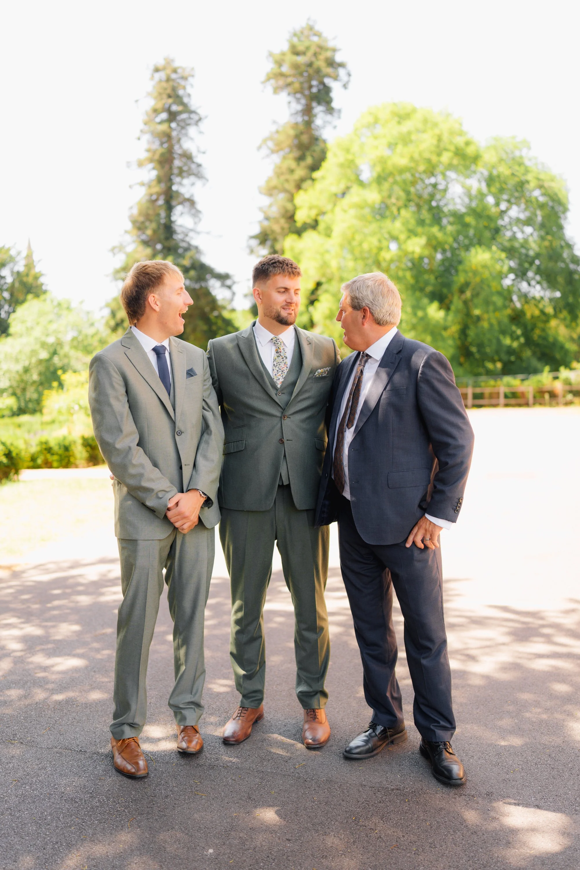 Three men in suits standing outdoors and engaged in conversation, with a background of trees and greenery.