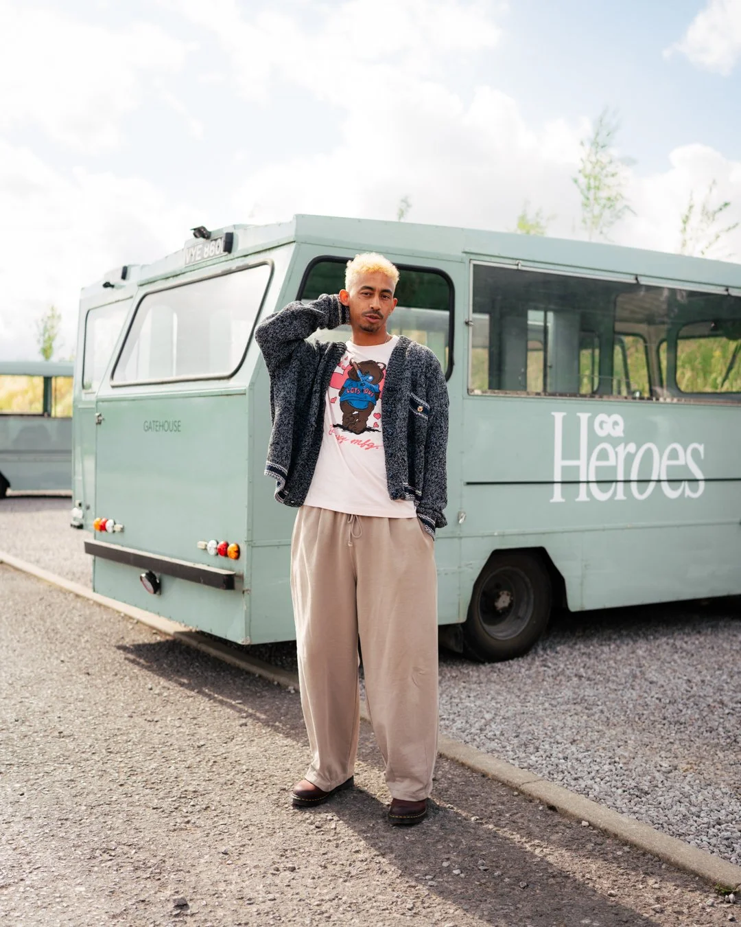 A young man with bleached blonde hair standing outdoors in front of a pale green, vintage-style bus with the words 'Heroes' on the side. He is dressed casually in beige loose-fitting pants, a graphic t-shirt, and a dark cardigan, with one hand touchi