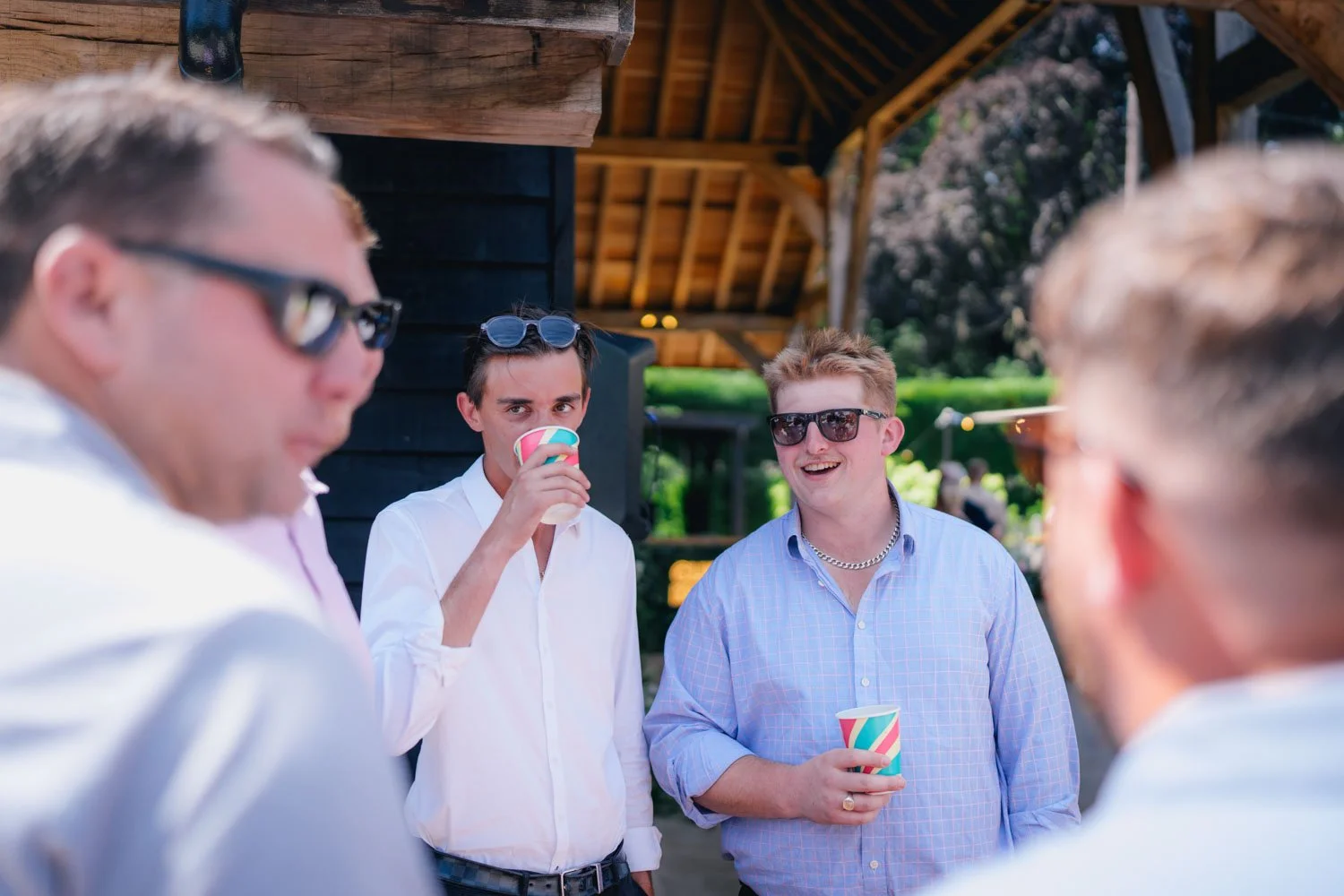 Group of young men enjoying drinks outdoors at a social gathering, with some wearing sunglasses and casual clothing, under a wooden shelter with greenery in the background.