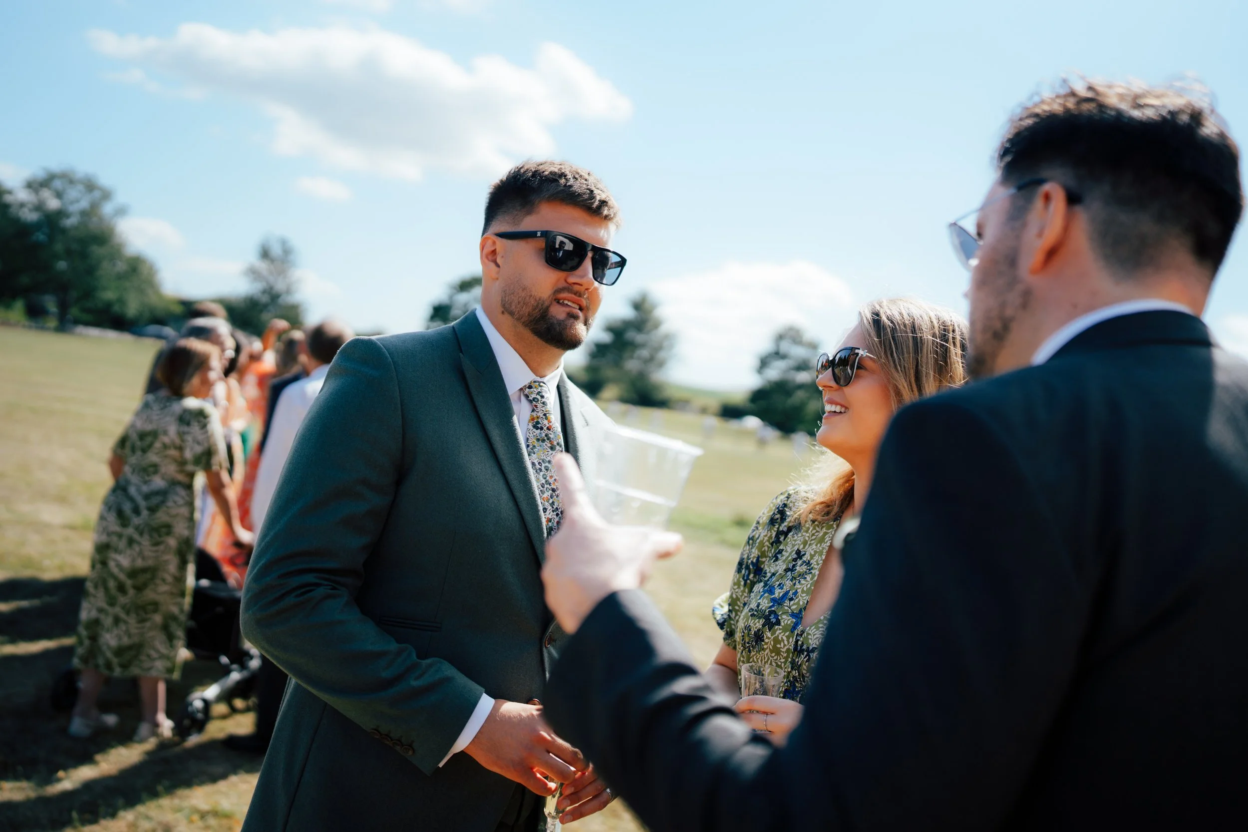 People at an outdoor social gathering, dressed in formal attire, some wearing sunglasses, engaging in conversation on a sunny day with a grassy field and trees in the background.