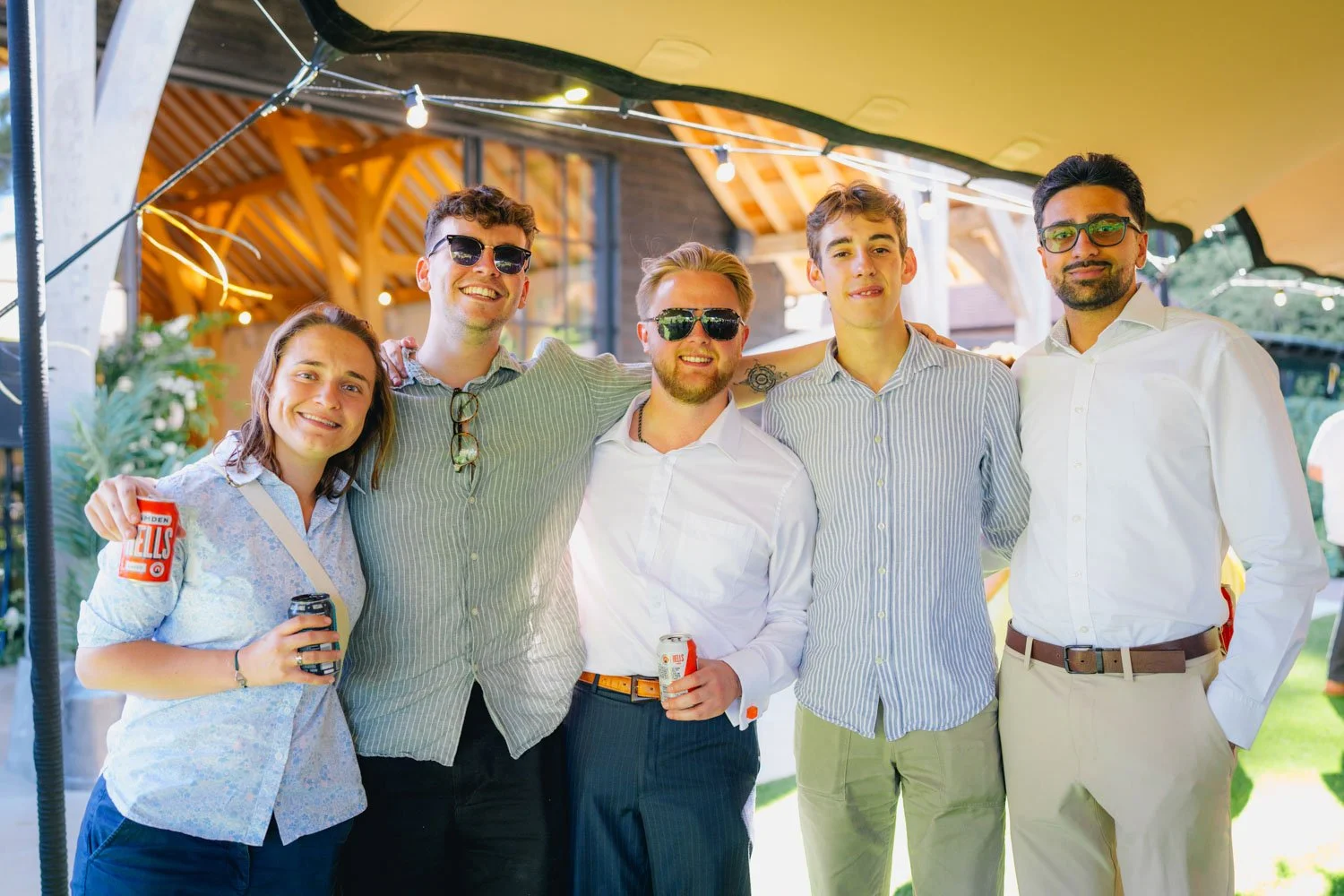 Group of five young adults smiling and posing for photo at outdoor gathering, some wearing sunglasses, holding drinks, under decorative string lights, with a building and greenery in the background.