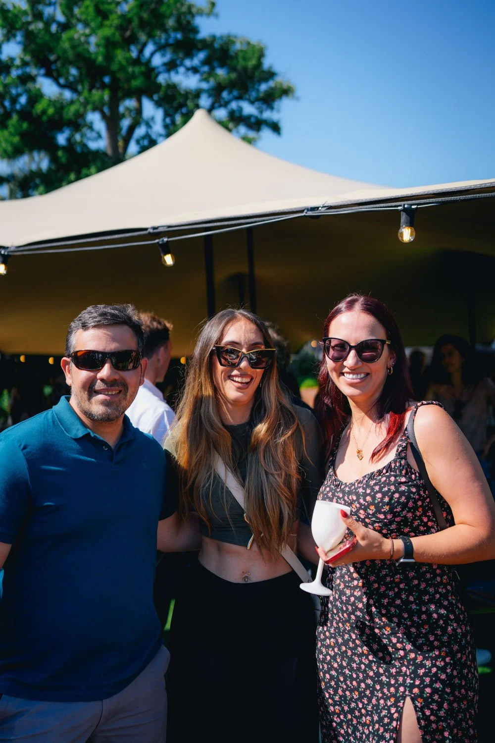 Three smiling people, two women and a man, standing outdoors during daytime at a sunny event, under a yellow tent, with trees in the background. The women are wearing sunglasses, with one holding a white drink in a champagne glass.