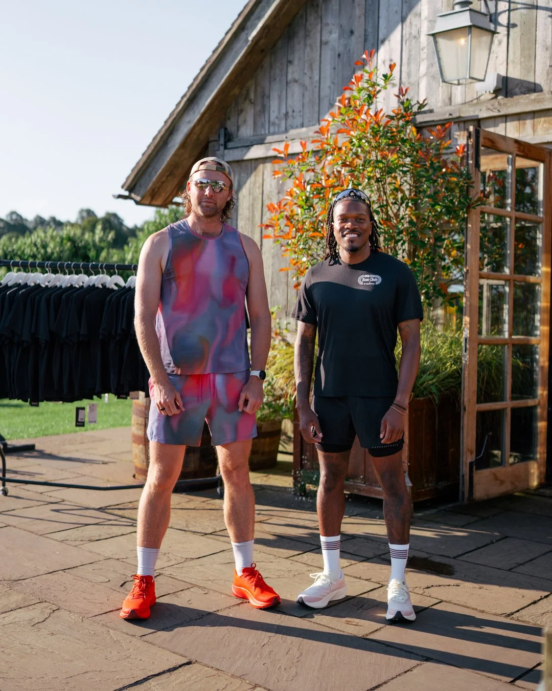 Two men standing outdoors in front of a rustic wooden building with a lush green background, both dressed in athletic wear, smiling at the camera.