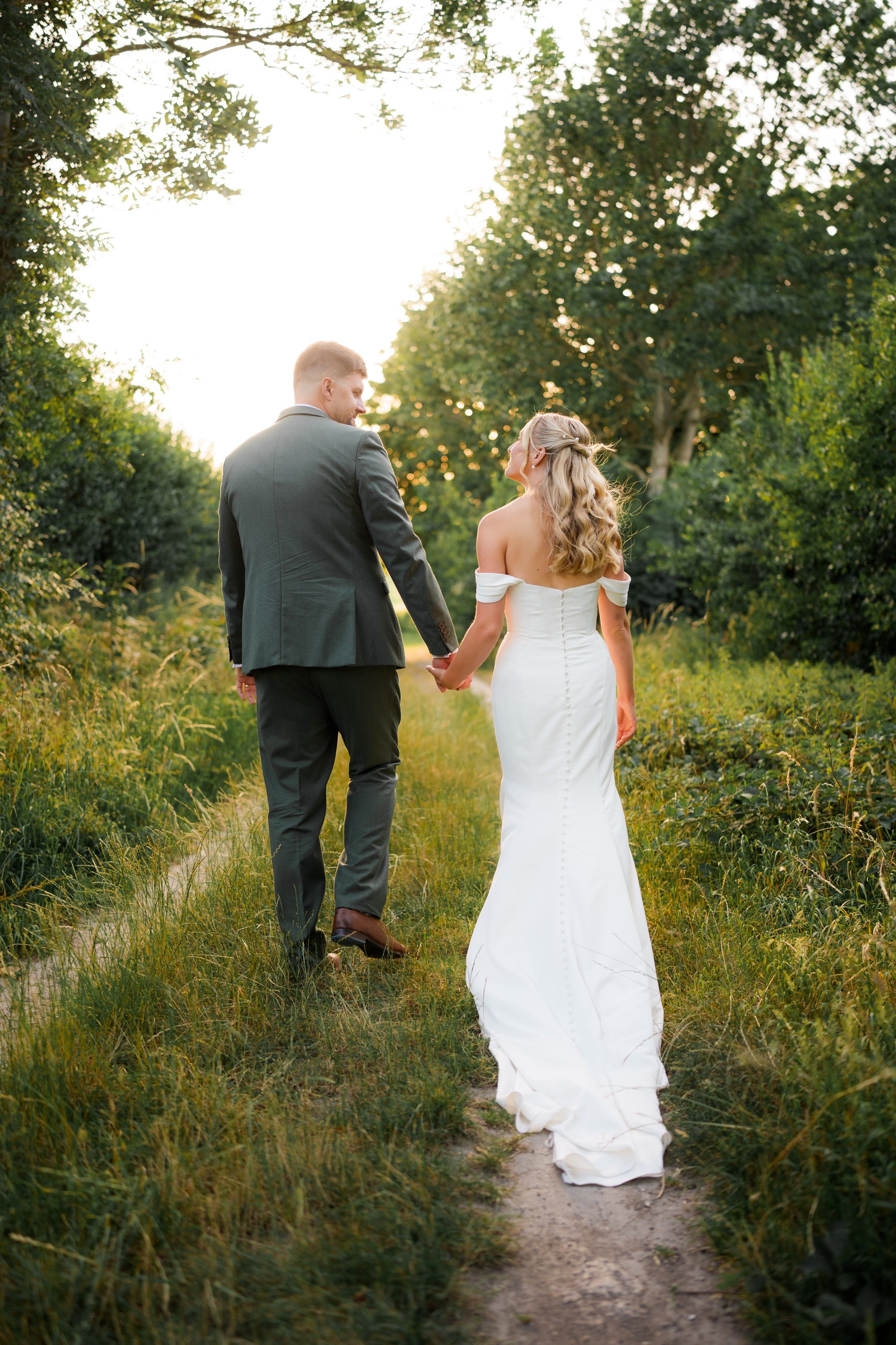 A bride and groom hold hands as they walk along a narrow dirt path through a lush green forest, with sunlight filtering through the trees.