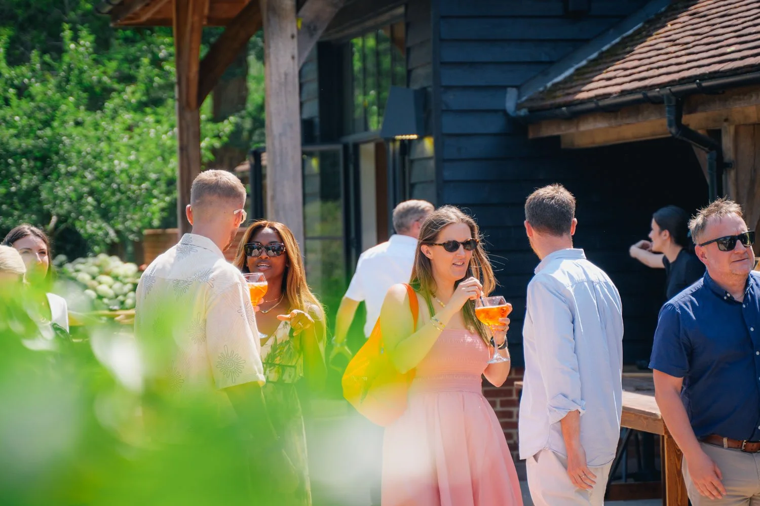 People socializing outdoors at a gathering, some holding drinks, near a dark wooden building with a brick foundation, surrounded by green trees on a sunny day.