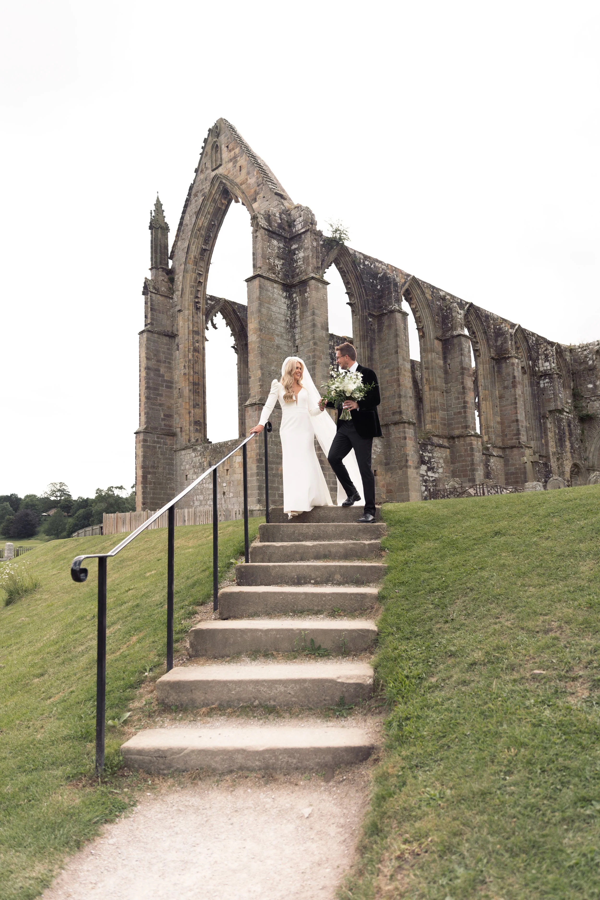 A bride and groom standing on steps outside of a historic stone ruin, exchanging vows or sharing an intimate moment, with the bride in a white wedding dress holding a bouquet, and the groom in a black tuxedo.