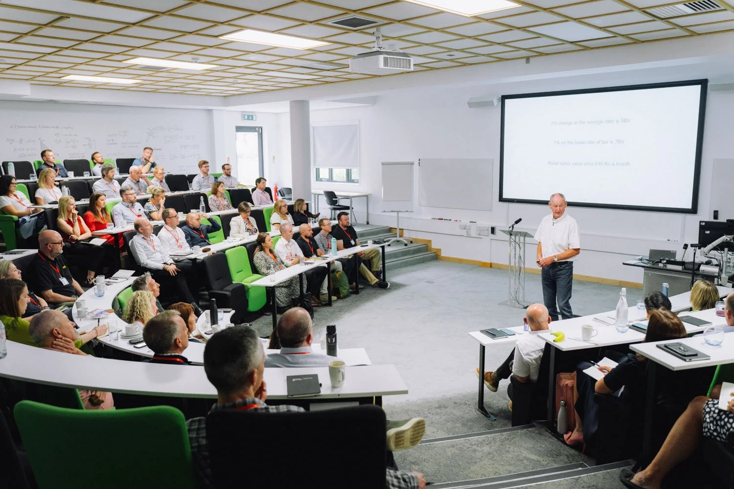 A presentation or lecture in a large, modern lecture hall with a diverse audience. The speaker, an elderly man, stands in front of a large screen with a PowerPoint slide. Attendees are seated in tiered rows, taking notes, and listening attentively.