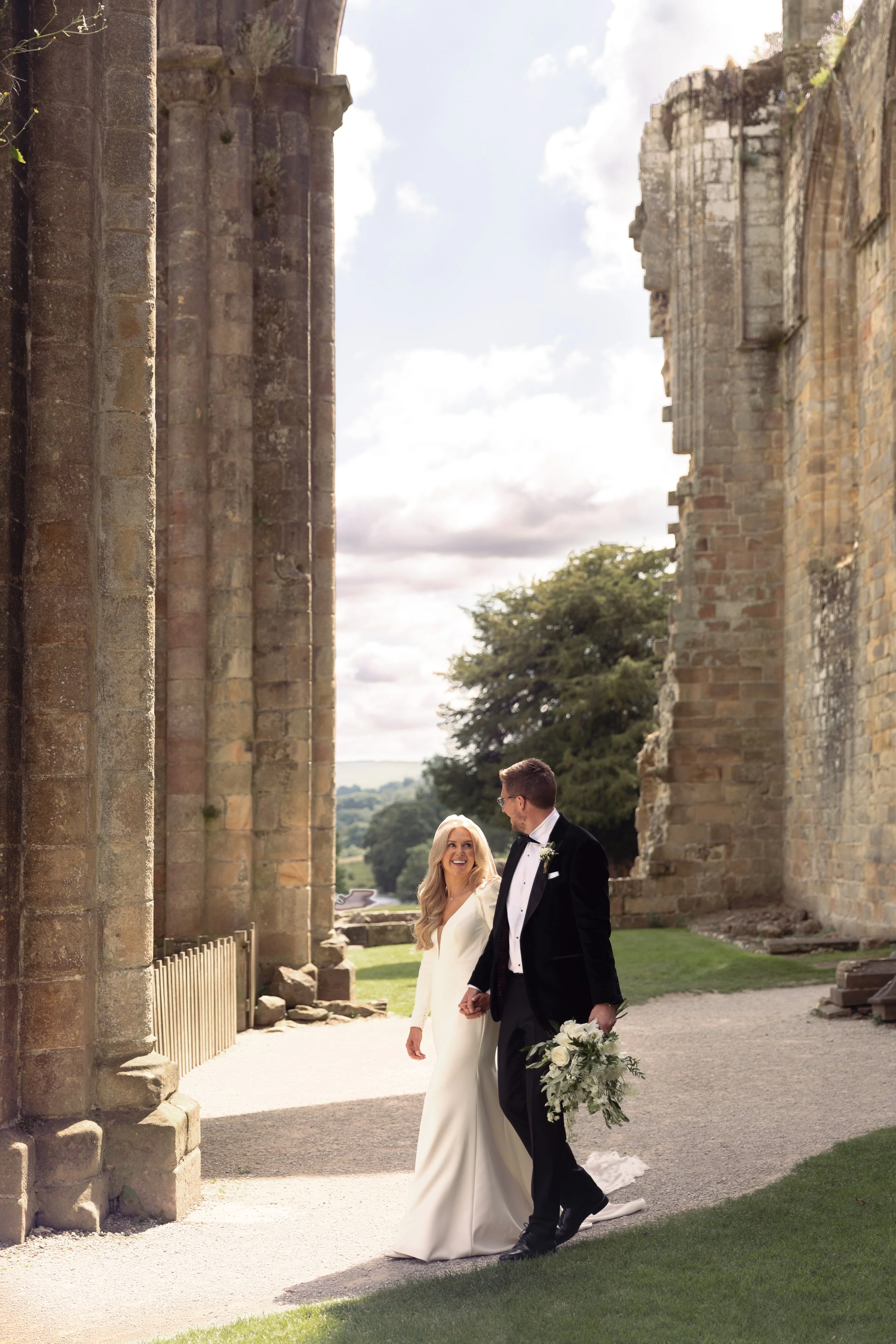 A bride and groom walking hand in hand outdoors, with the bride smiling at the groom, near stone ruins and large trees on a partly cloudy day.