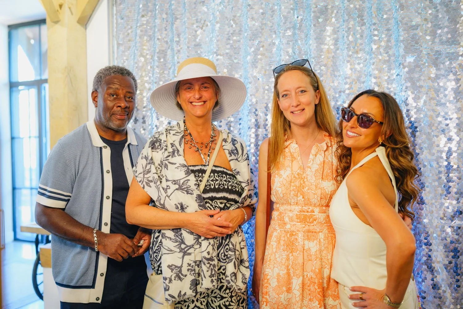 Four adults posing together indoors, standing in front of a shiny, sequin-covered backdrop. The group includes an older man and women wearing summery, casual attire, with the women on the right wearing sunglasses and smiling.