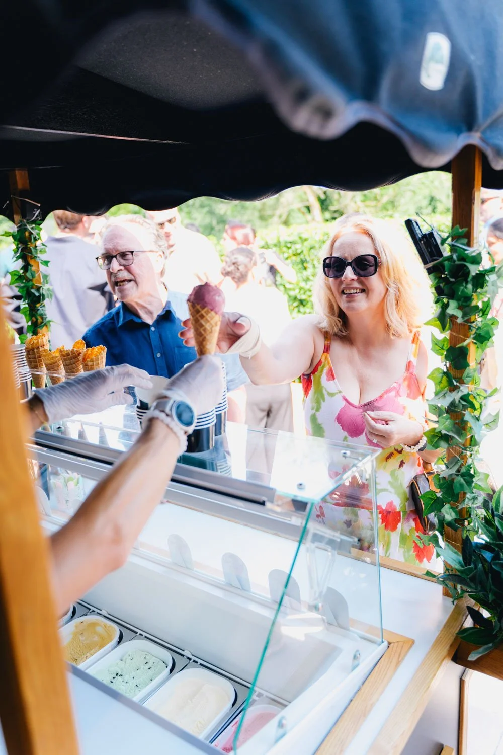 A woman in a floral dress and sunglasses is happy as she receives a cone of purple ice cream from an ice cream vendor at an outdoor stand, with a man in a blue shirt behind her. Other customers are in the background.
