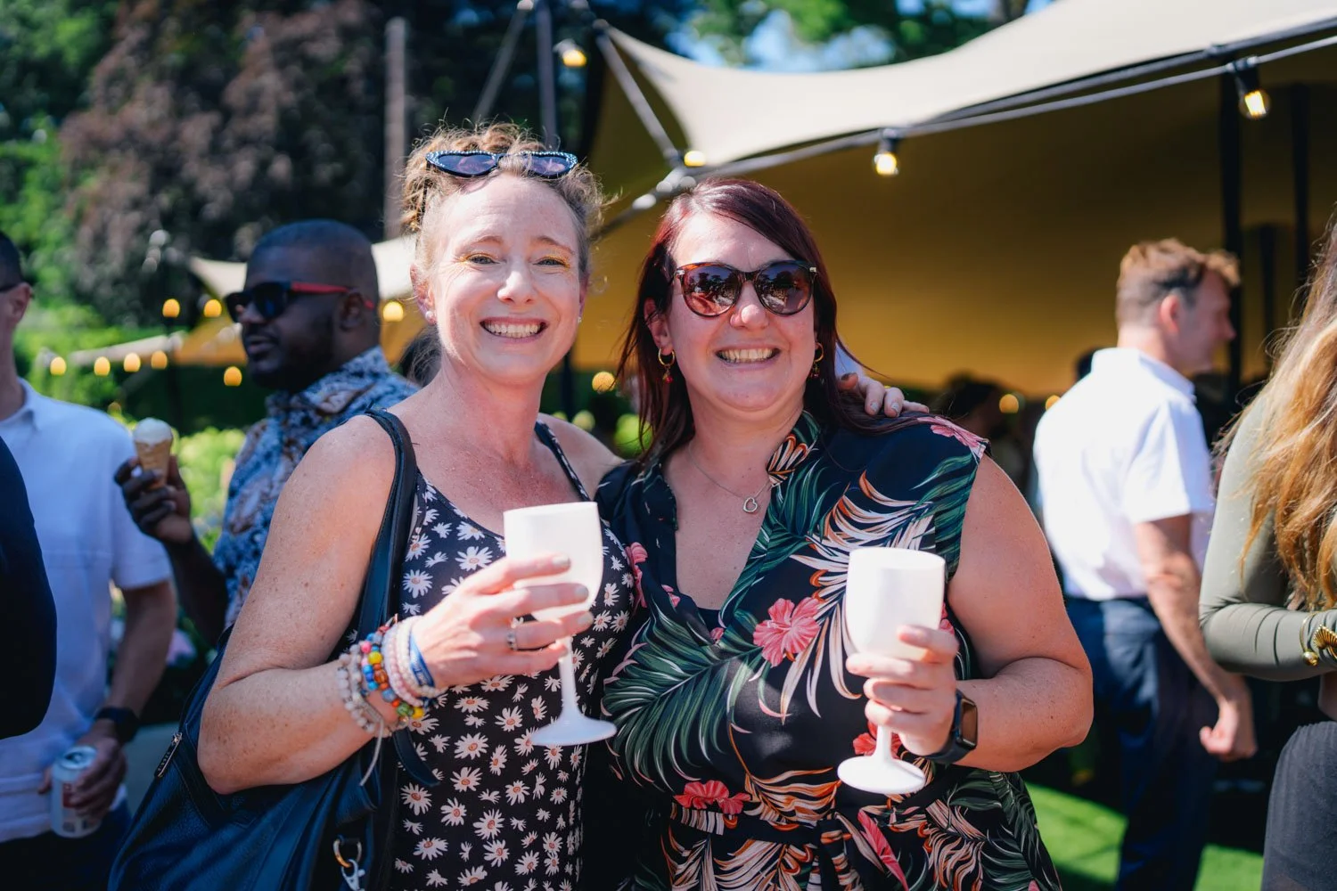 Two women smiling and holding drinks at an outdoor event, with other guests in the background.