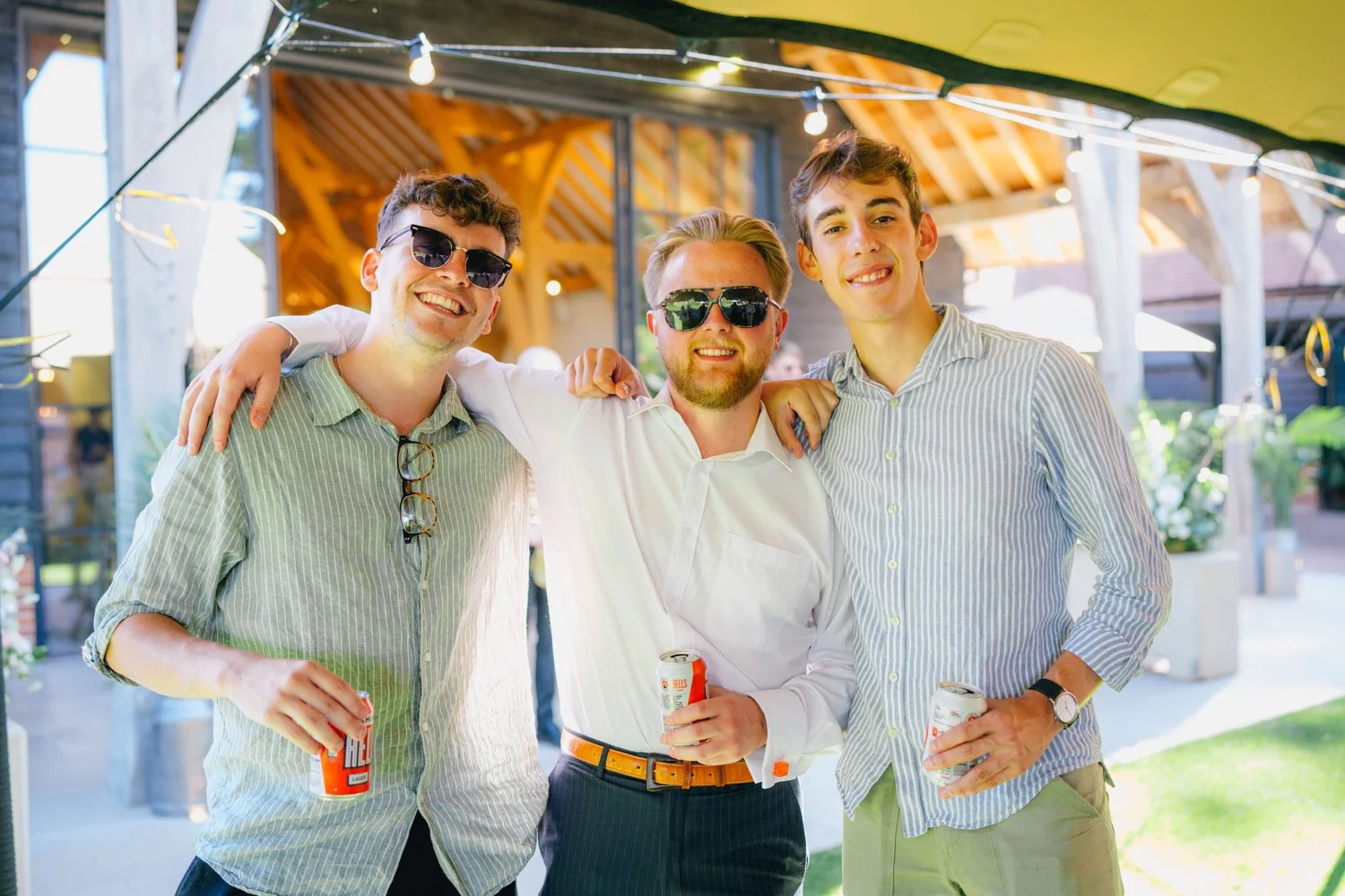 Three young men smiling and enjoying drinks together at an outdoor celebration under a canopy with string lights.