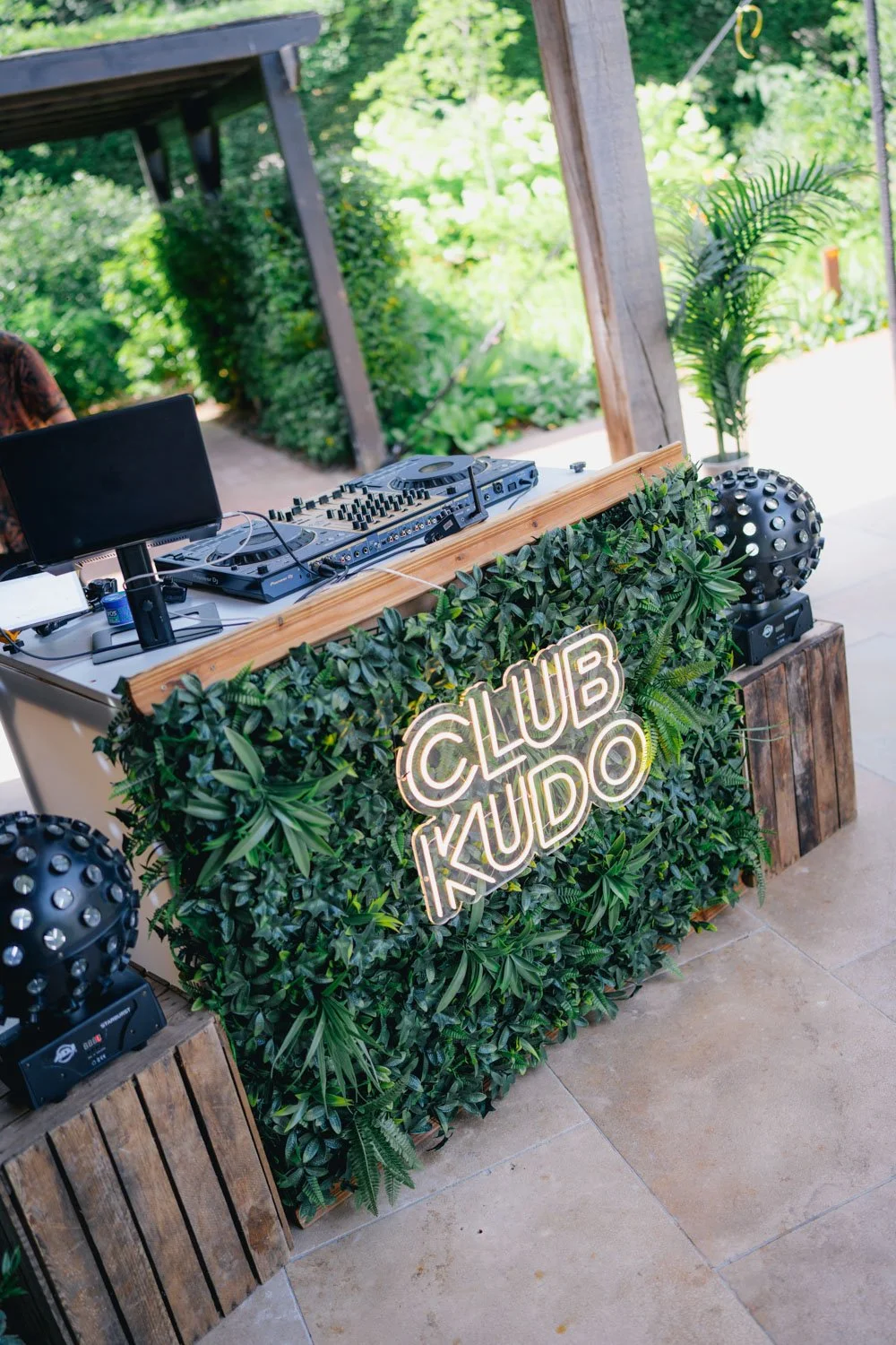 DJ setup with a neon sign reading 'Club Kudo' on a table decorated with green foliage, with speakers and DJ equipment outdoors.
