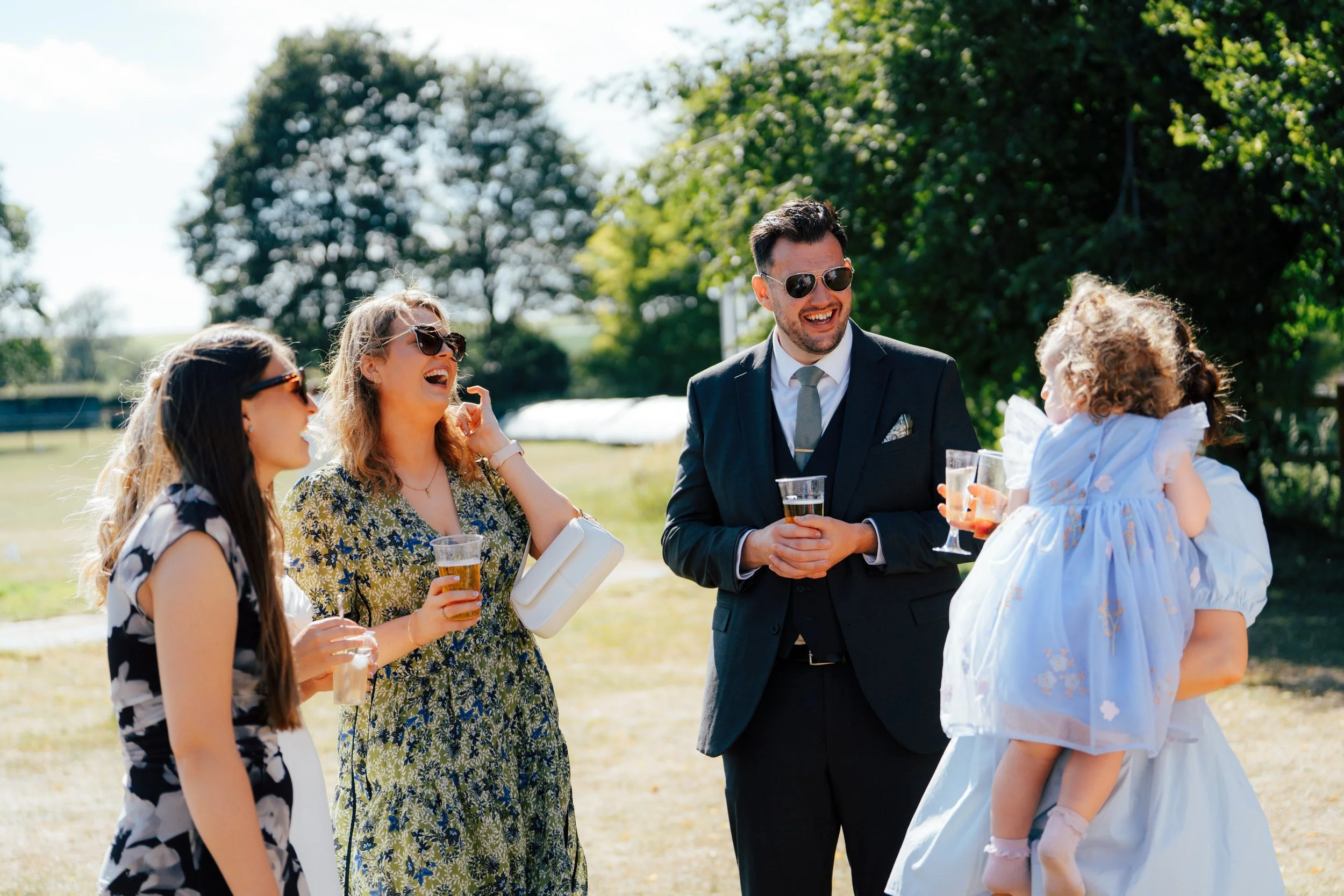 Group of people outdoors enjoying drinks, including a man in a suit and sunglasses, women wearing sunglasses, and a little girl in a dress, with trees and green landscape in the background.
