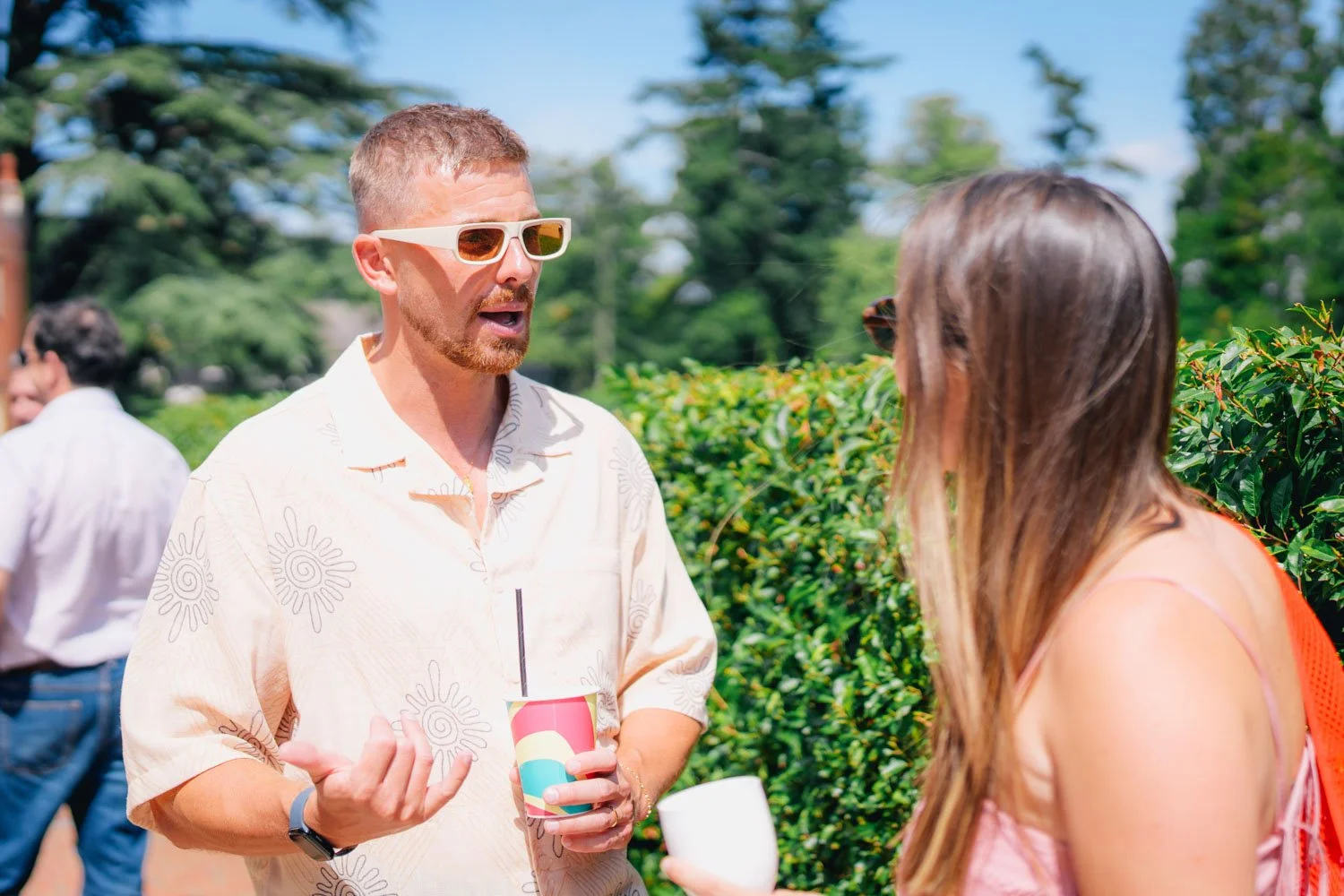 Two people, a man with short hair, sunglasses, and a beard in a light patterned shirt, and a woman with long brown hair in a pink dress, are having a conversation outdoors on a sunny day, with greenery and other people in the background.