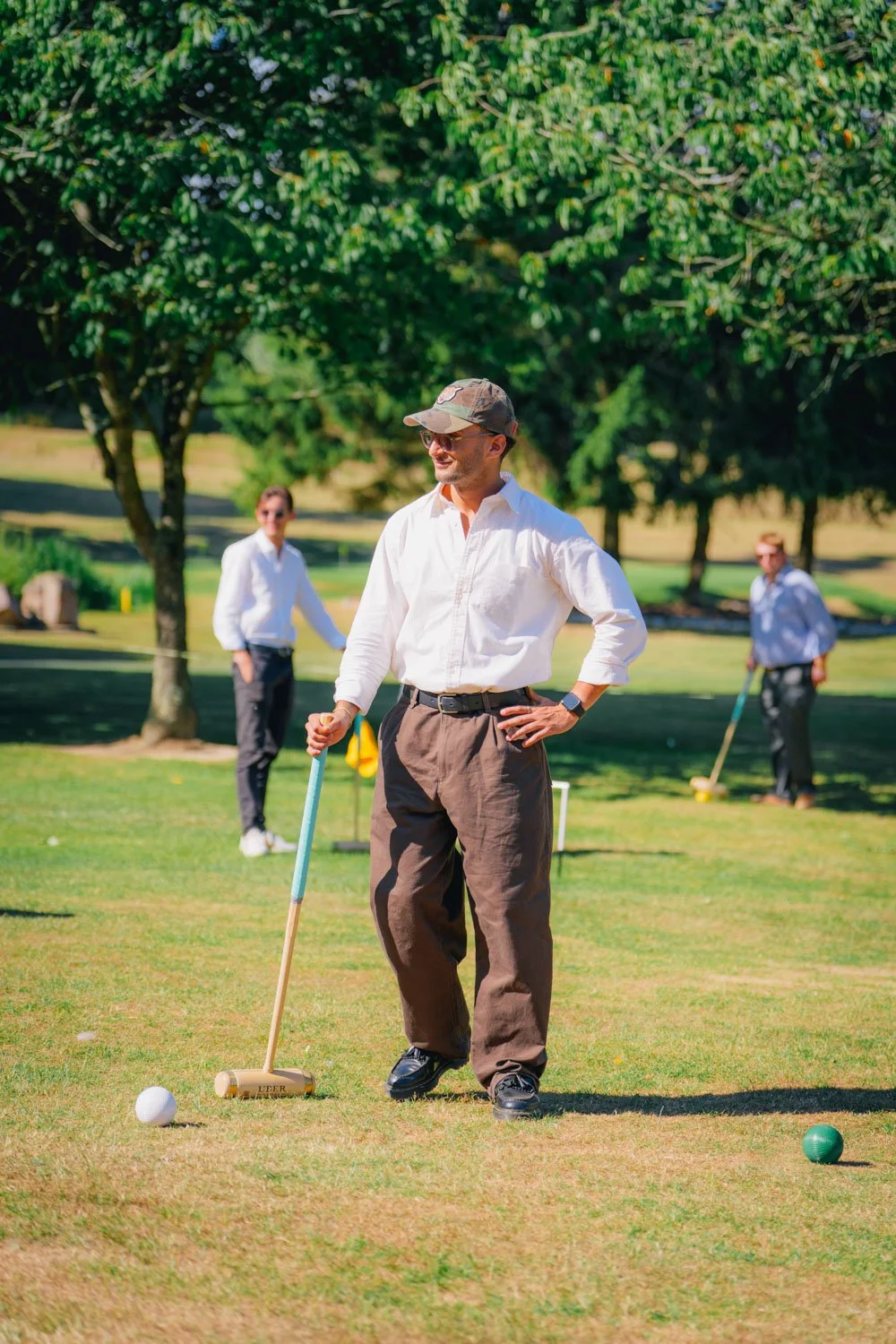 A man in a white shirt and brown pants standing on a golf course, holding a chipping putter, with other people playing in the background.
