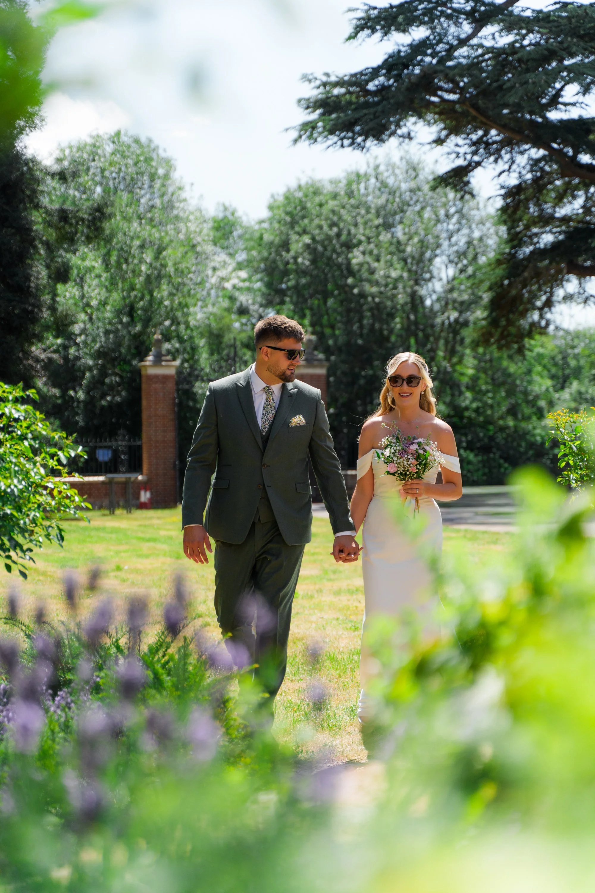 A bride and groom walking outdoors in a garden on their wedding day, holding hands, with the groom dressed in a suit and the bride in a wedding dress holding a bouquet of flowers.