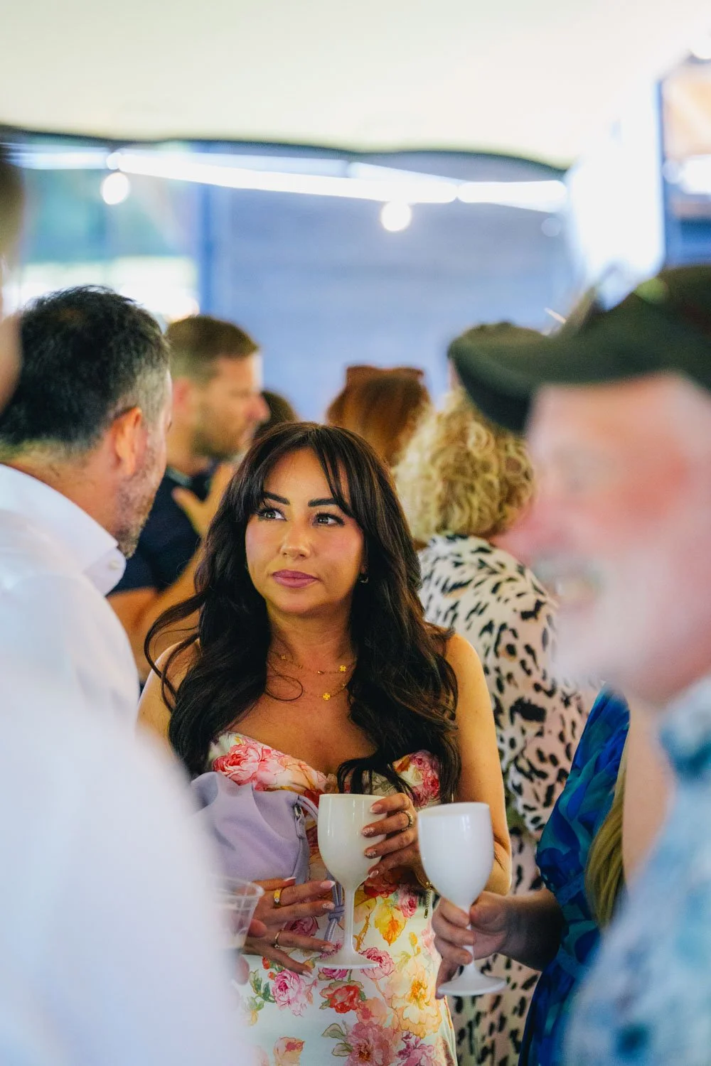 People at a social gathering, with a woman in a floral dress holding a white wine glass, engaged in conversation in a bright indoor setting.