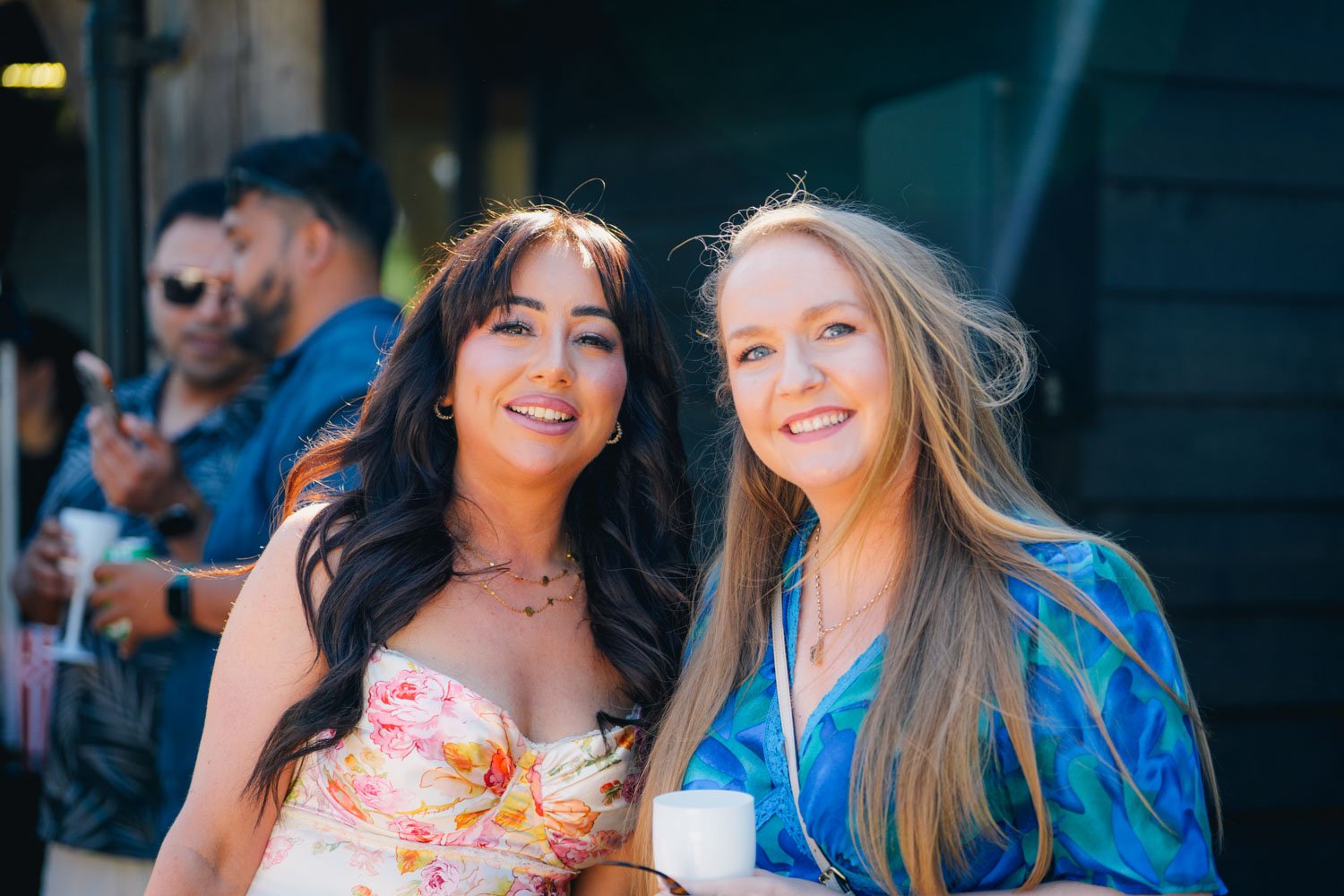 Two women smiling and posing outdoors at a social gathering, with other people in the background.