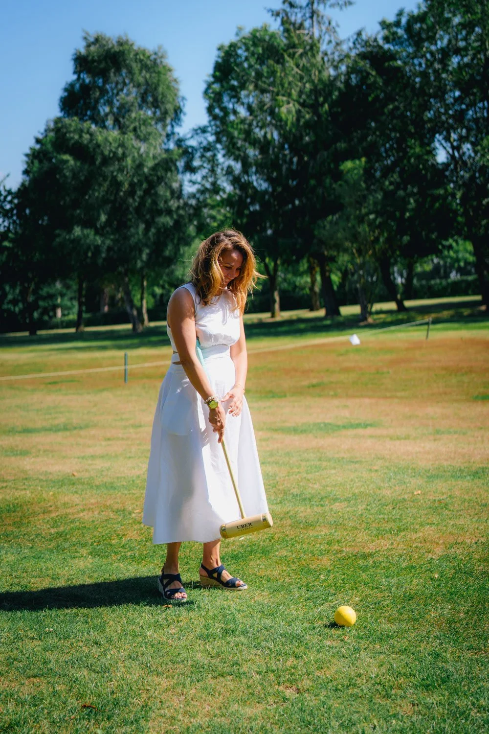 Woman in white dress playing croquet on a grassy field with trees in the background.